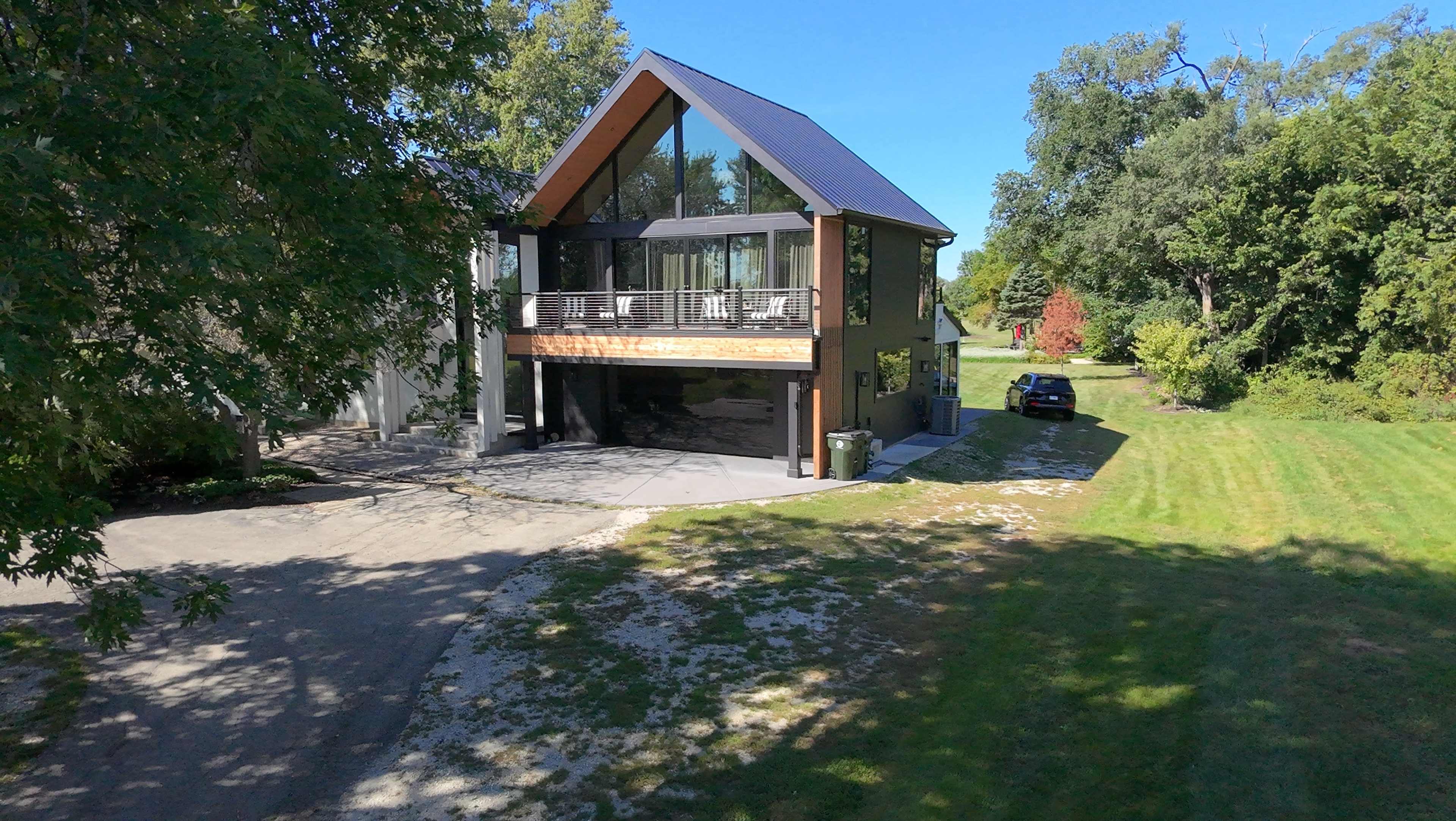 A modern house with a triangular roof and large windows is situated next to a driveway and grassy area, surrounded by trees.