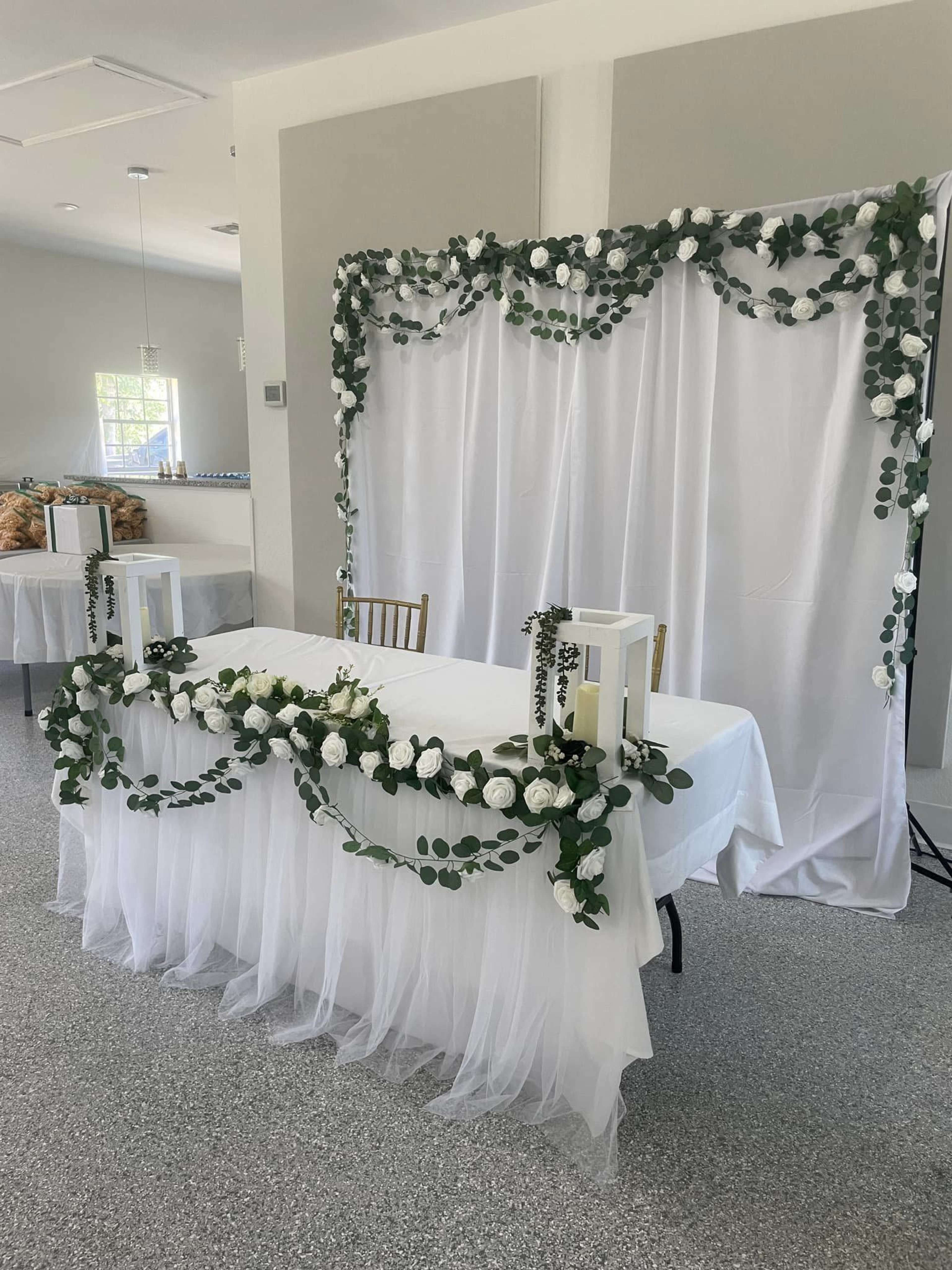 A decorated table is adorned with white flowers and greenery, set against a backdrop of draped white fabric.