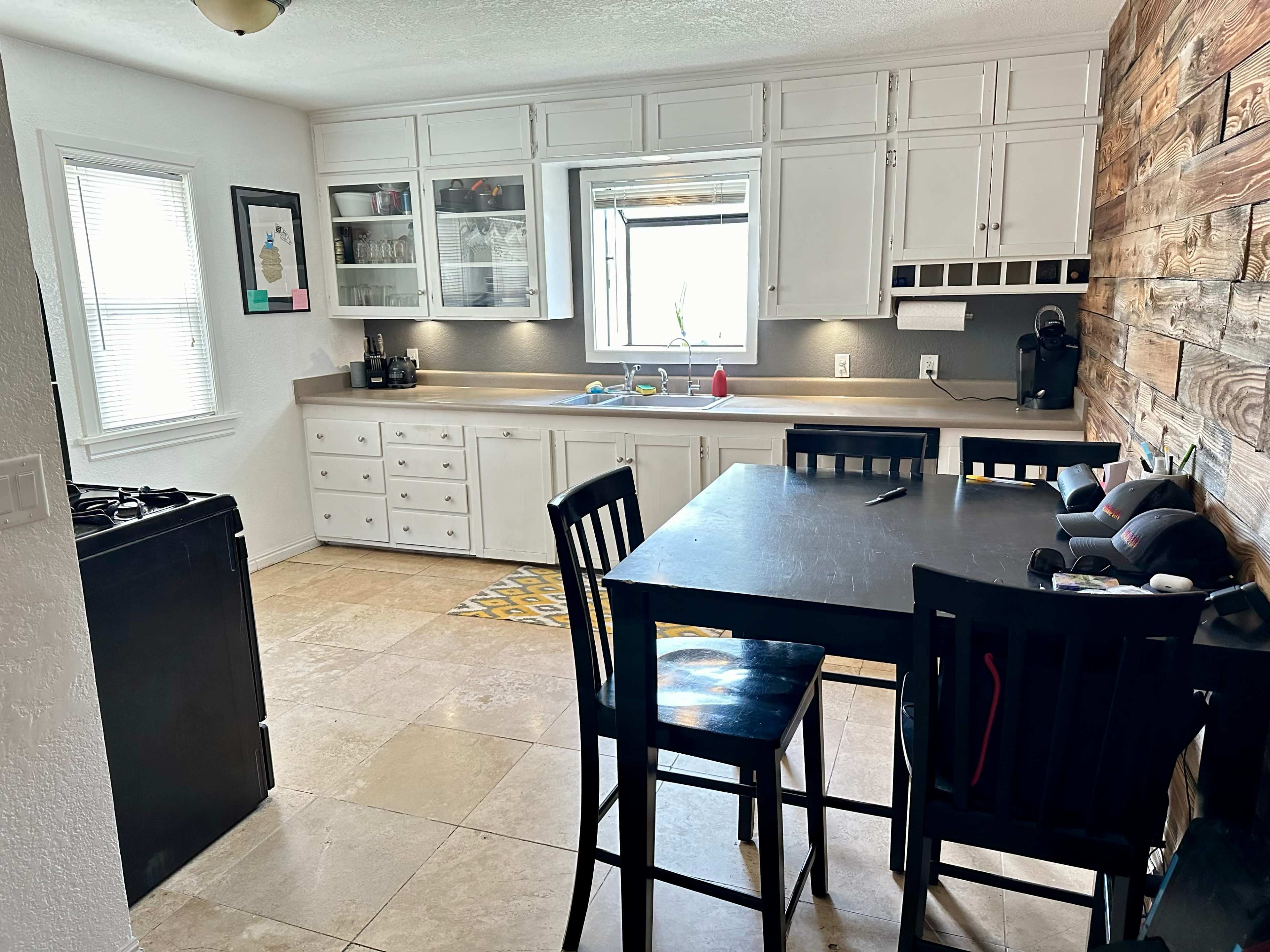 A kitchen with white cabinetry, a window above the sink, a black table with chairs, and tile flooring.