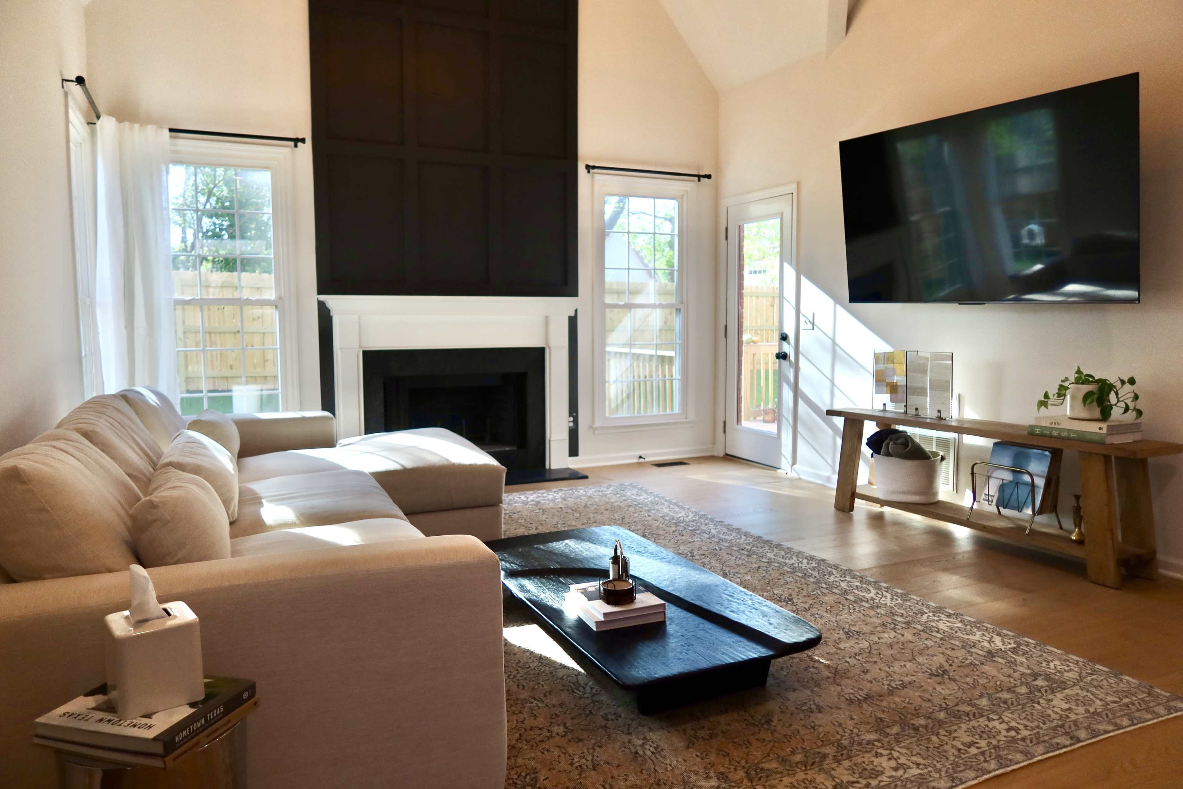 The image shows a bright living room with a white sectional sofa, a large black fireplace, a TV mounted on the wall, and sliding glass doors leading to a patio.