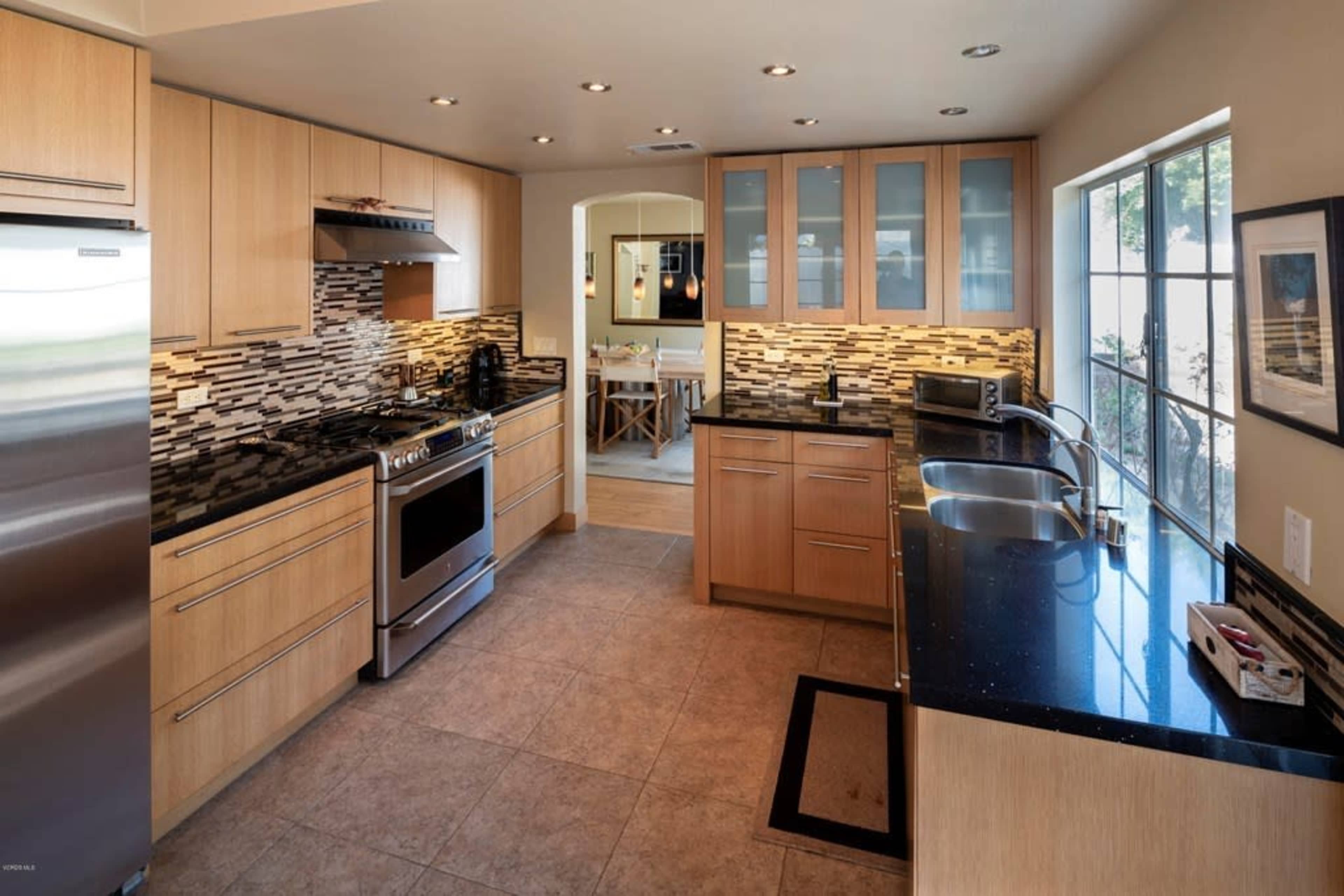 A modern kitchen with wooden cabinetry, black countertops, a stainless steel stove, and a window overlooking a dining area.