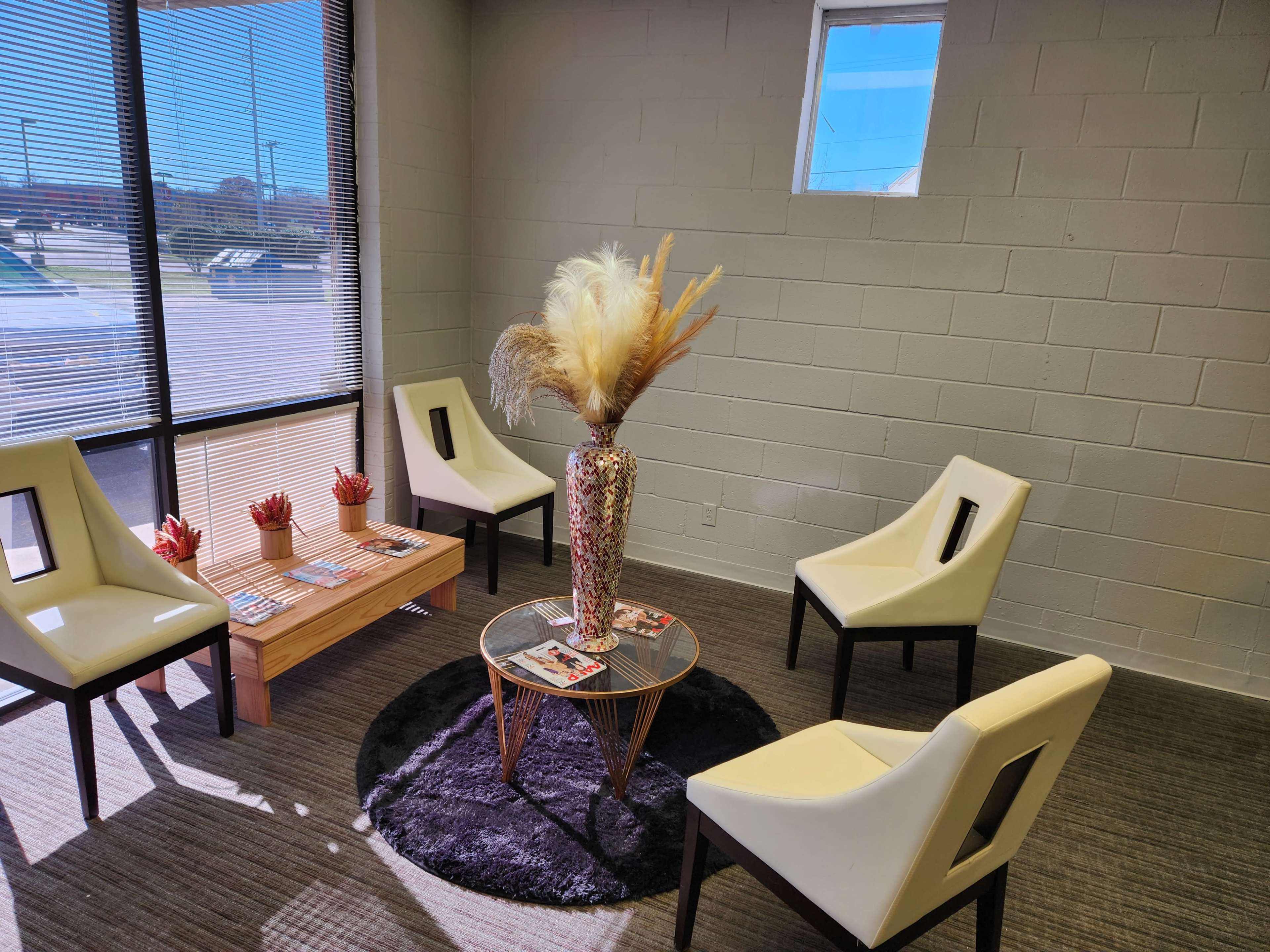A waiting area with four chairs arranged around a coffee table, featuring a vase with dried plants and magazines on the table.