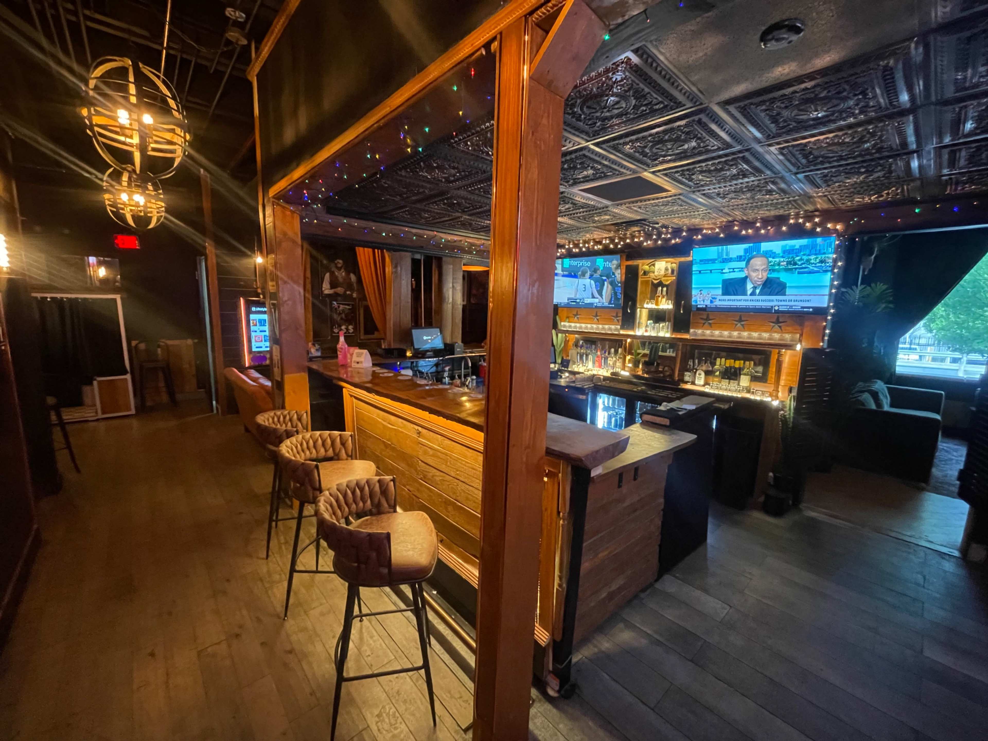 The image shows a dimly lit bar area with wooden accents, a TV displaying news, and a line of high stools facing the bar counter.
