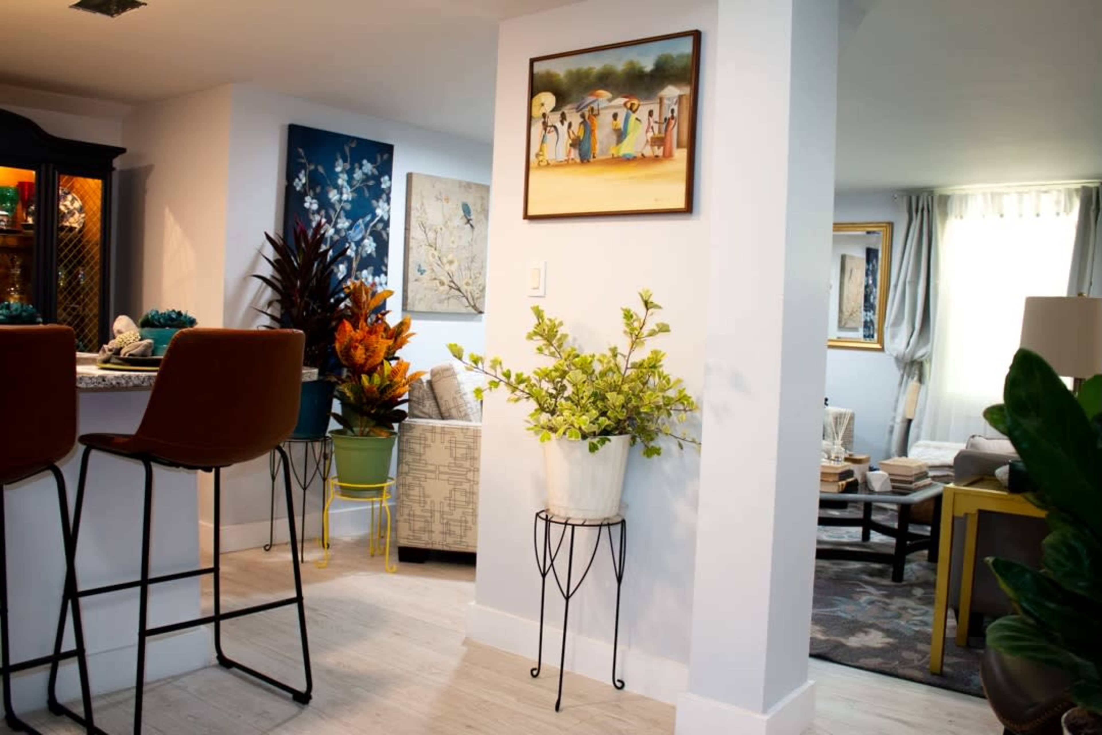 The image shows a bright, modern living space featuring a bar area with stools, decorative plants, and a framed painting on the wall.