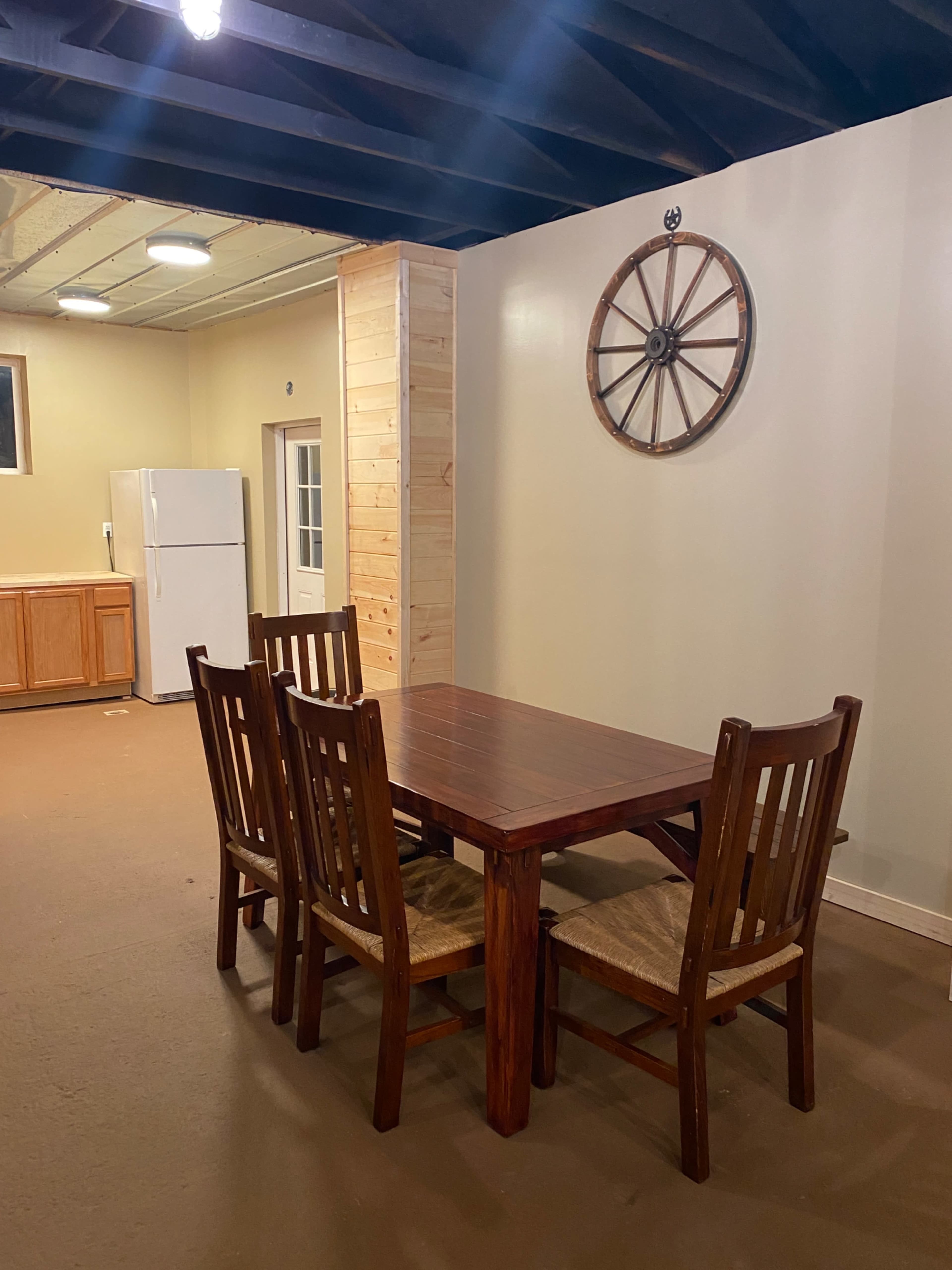 A wooden dining table with four chairs is set in a spacious room featuring a wheel decoration on the wall and a refrigerator in the background.