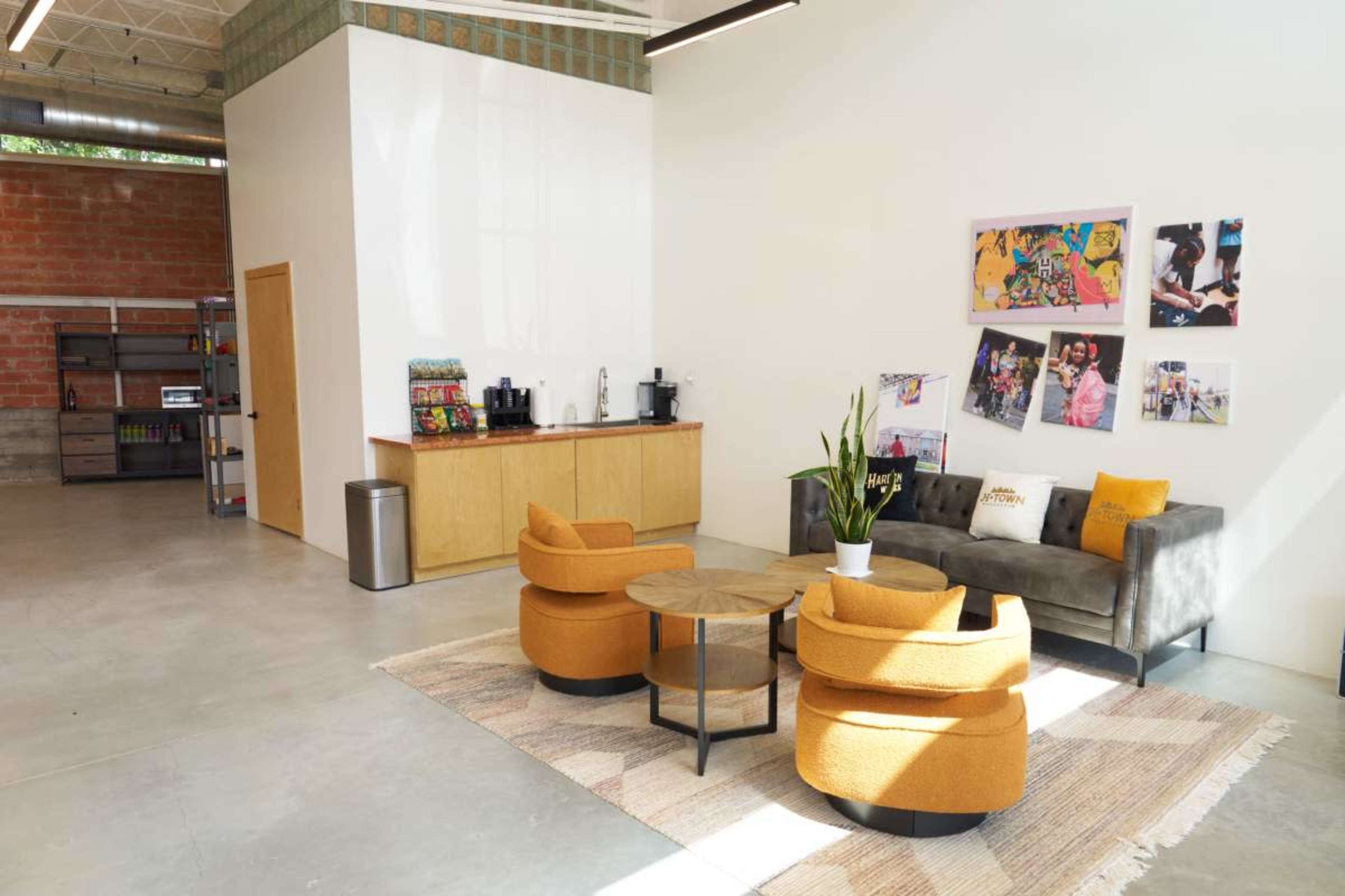 The image shows a modern lounge area with a grey sofa and two orange chairs, a round coffee table, a rug, and colorful artwork on the wall.