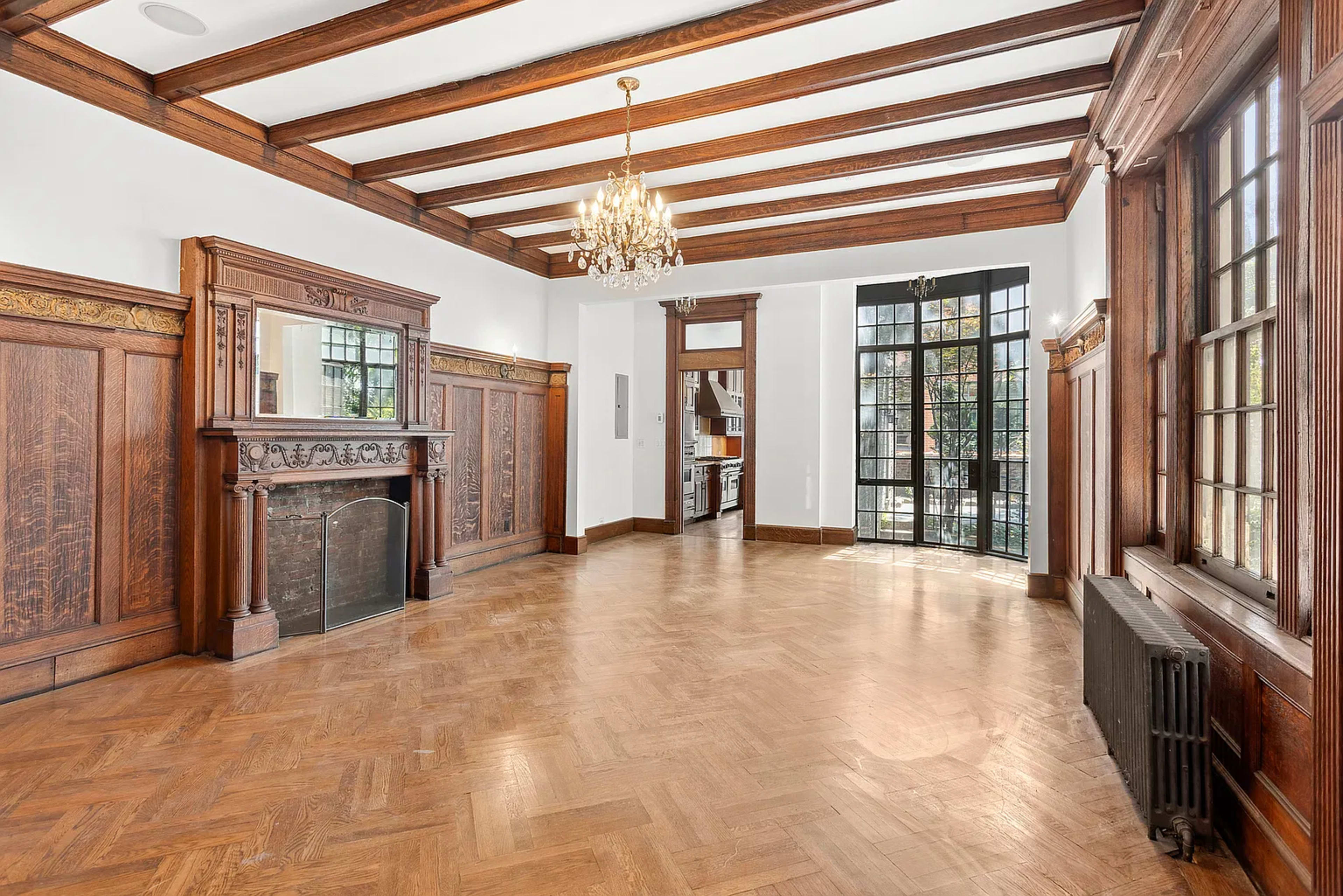The image shows a spacious interior room featuring wooden beams on the ceiling, a decorative fireplace, and large windows that illuminate the space.