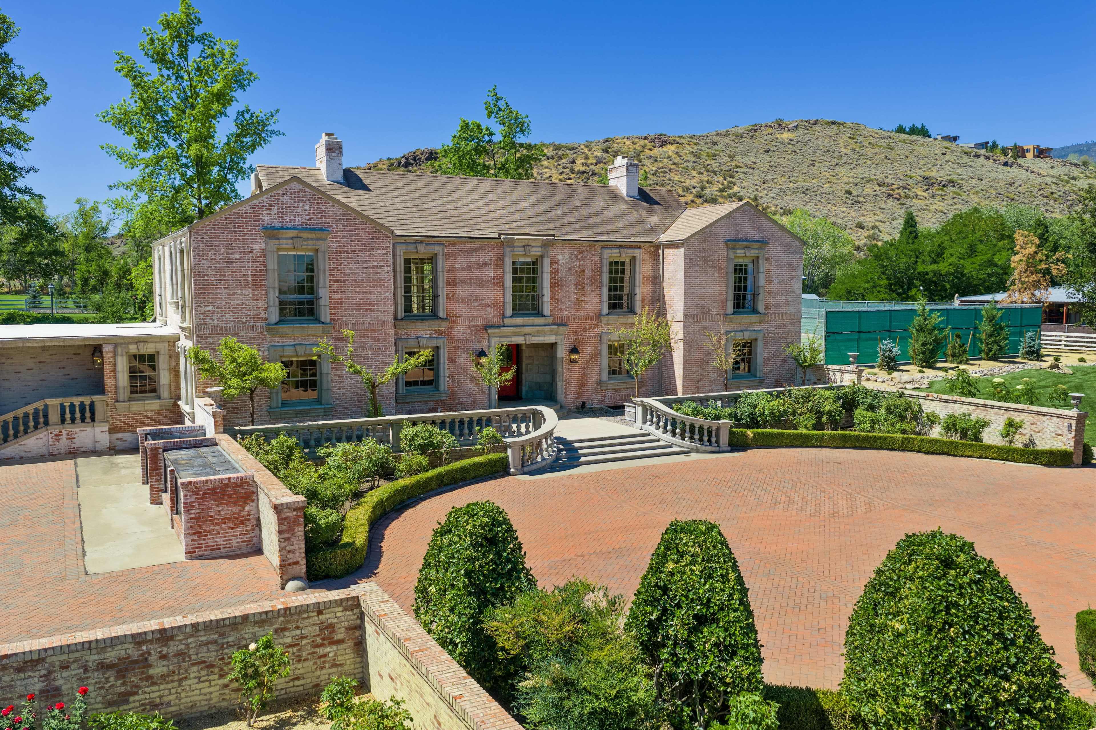 The image shows a large brick mansion surrounded by manicured gardens and trees, with a stone pathway leading to the entrance.