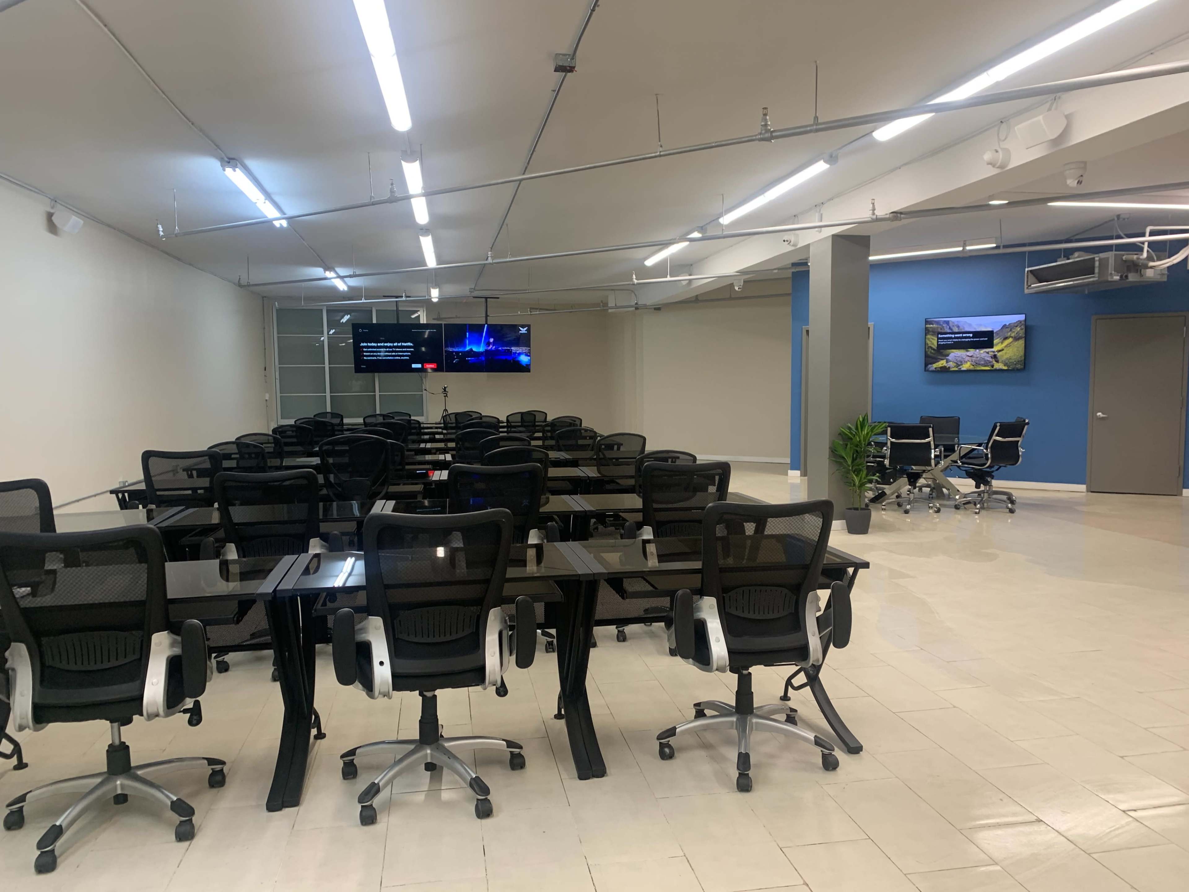 The image shows a modern conference room with multiple black ergonomic chairs arranged around large tables, two screens mounted on the walls, and a blue accent wall.