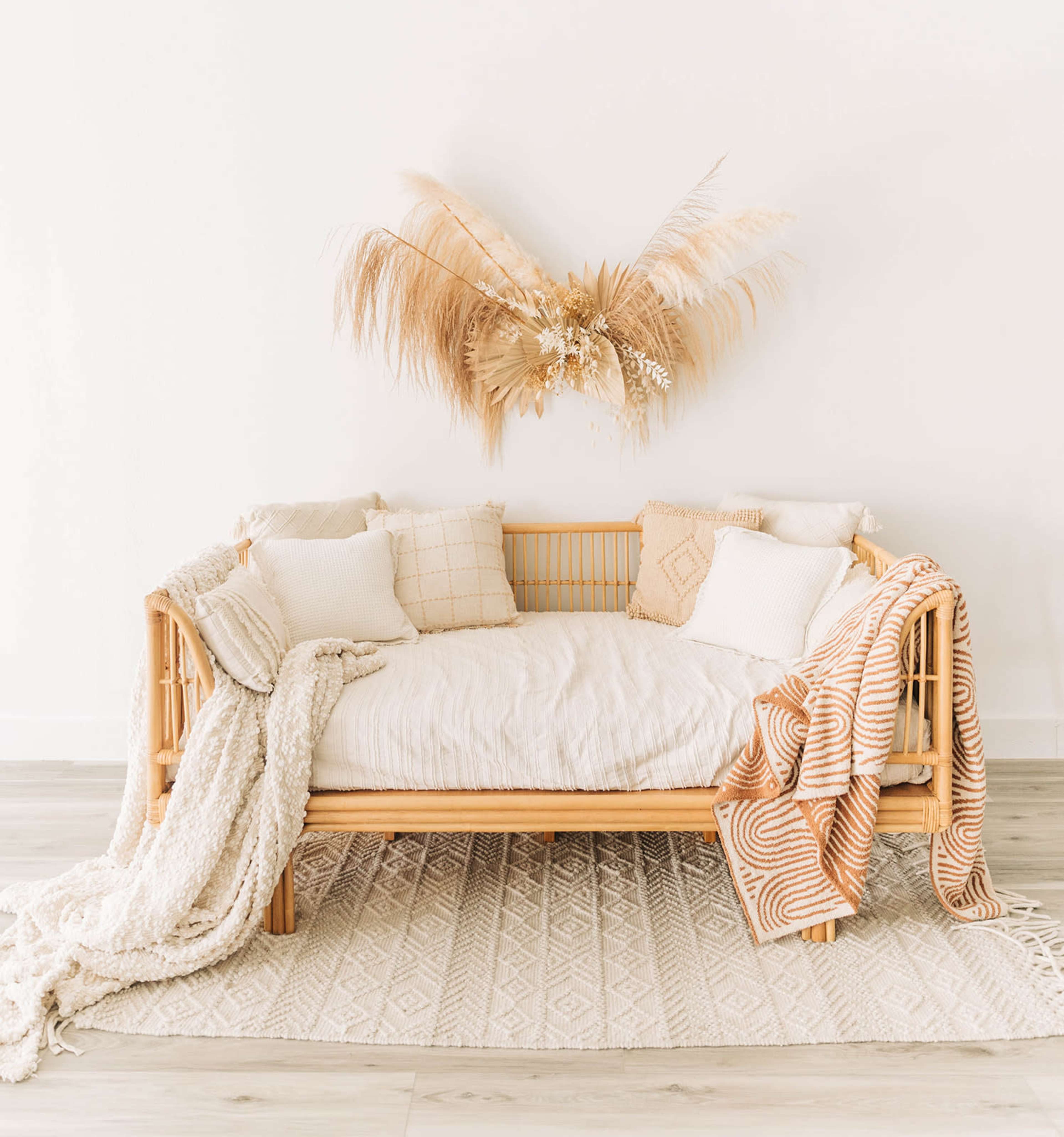 A rattan daybed is adorned with various cushions and blankets, positioned against a white wall with a decorative arrangement of dried grasses above it.