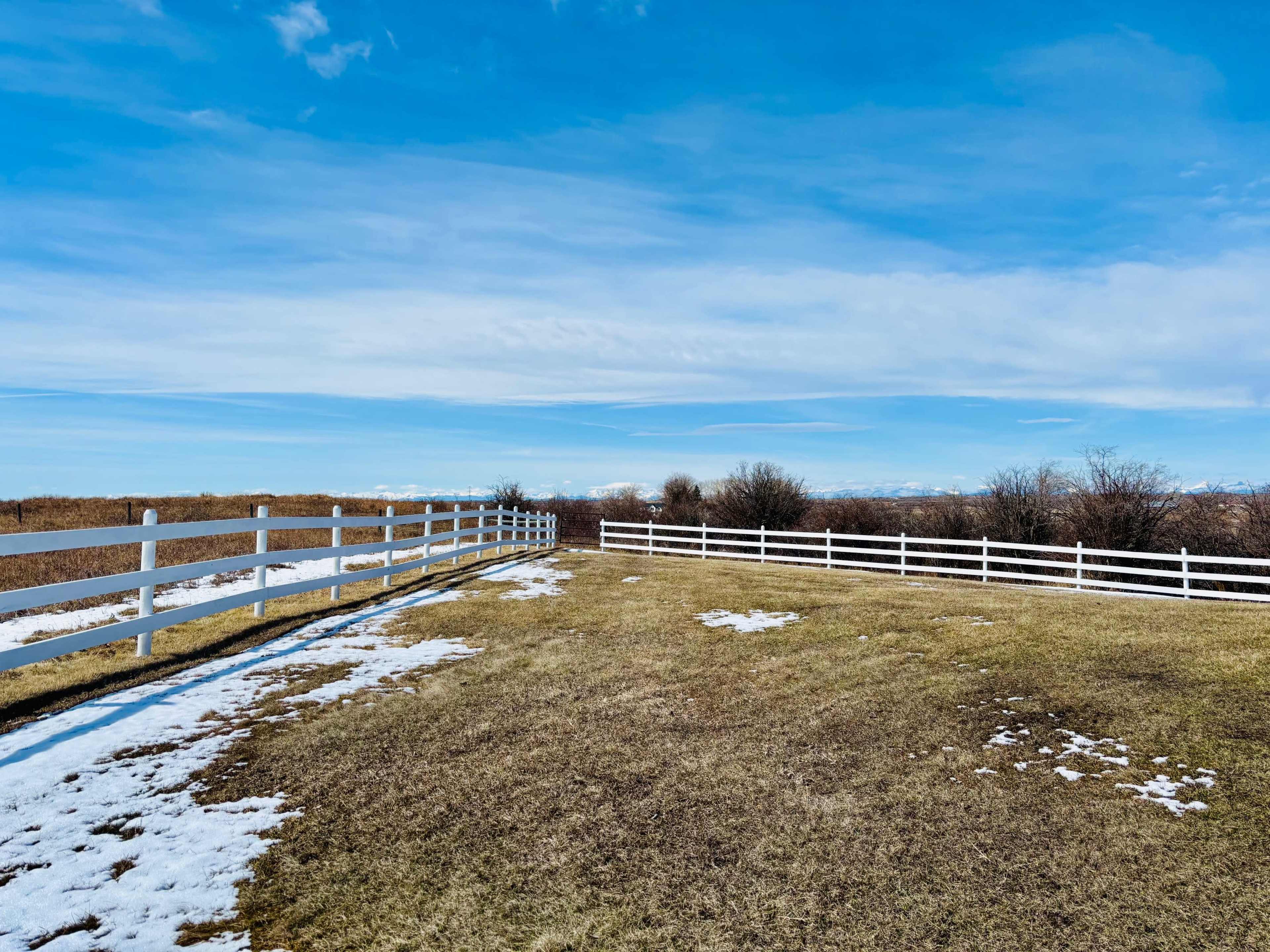A white fenced area with patches of snow and brown grass under a blue sky.