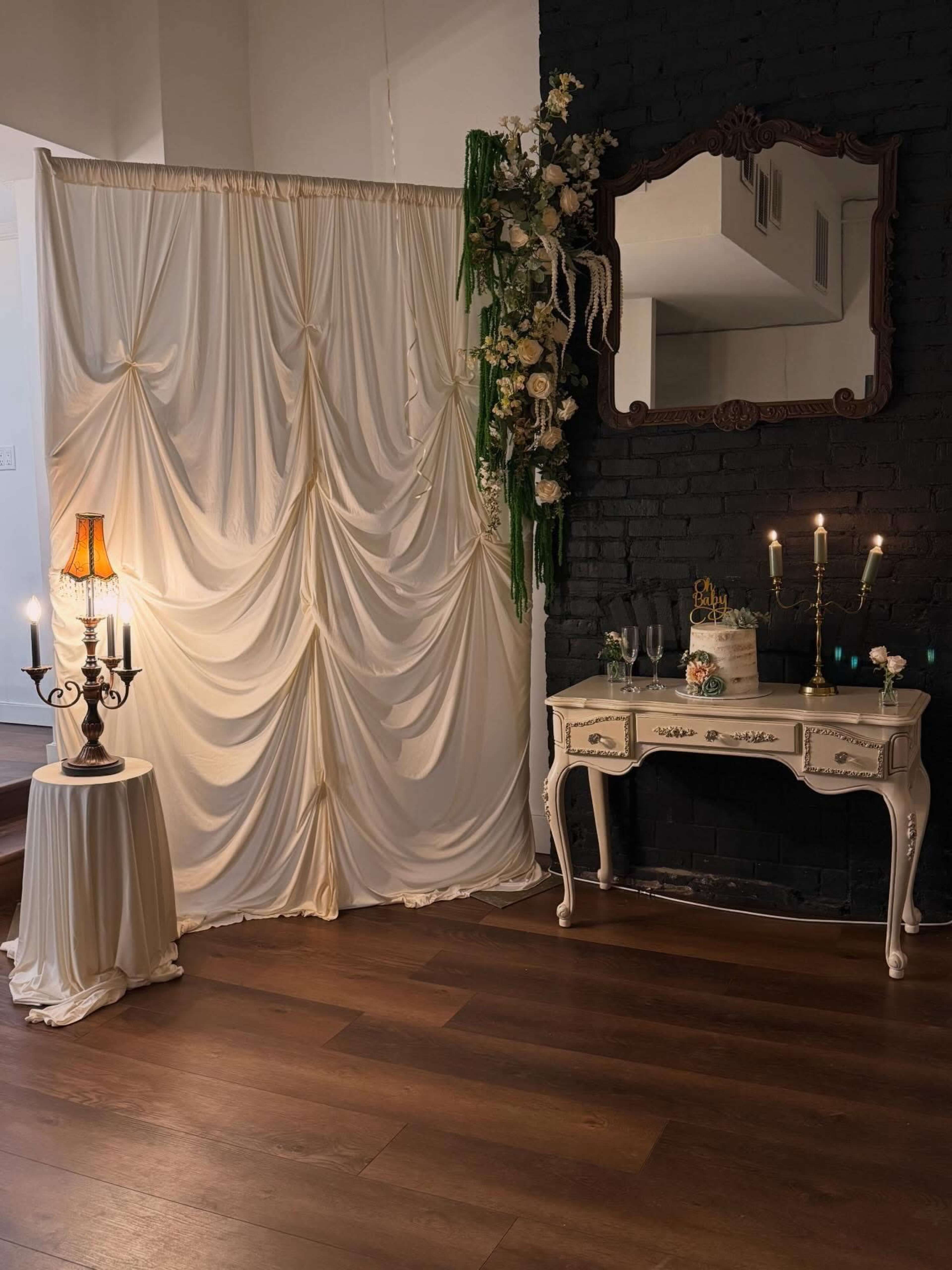 The scene features an elegantly decorated corner with a white draped curtain, a vintage mirror, a decorated table with candles and glassware, and a standing lamp.
