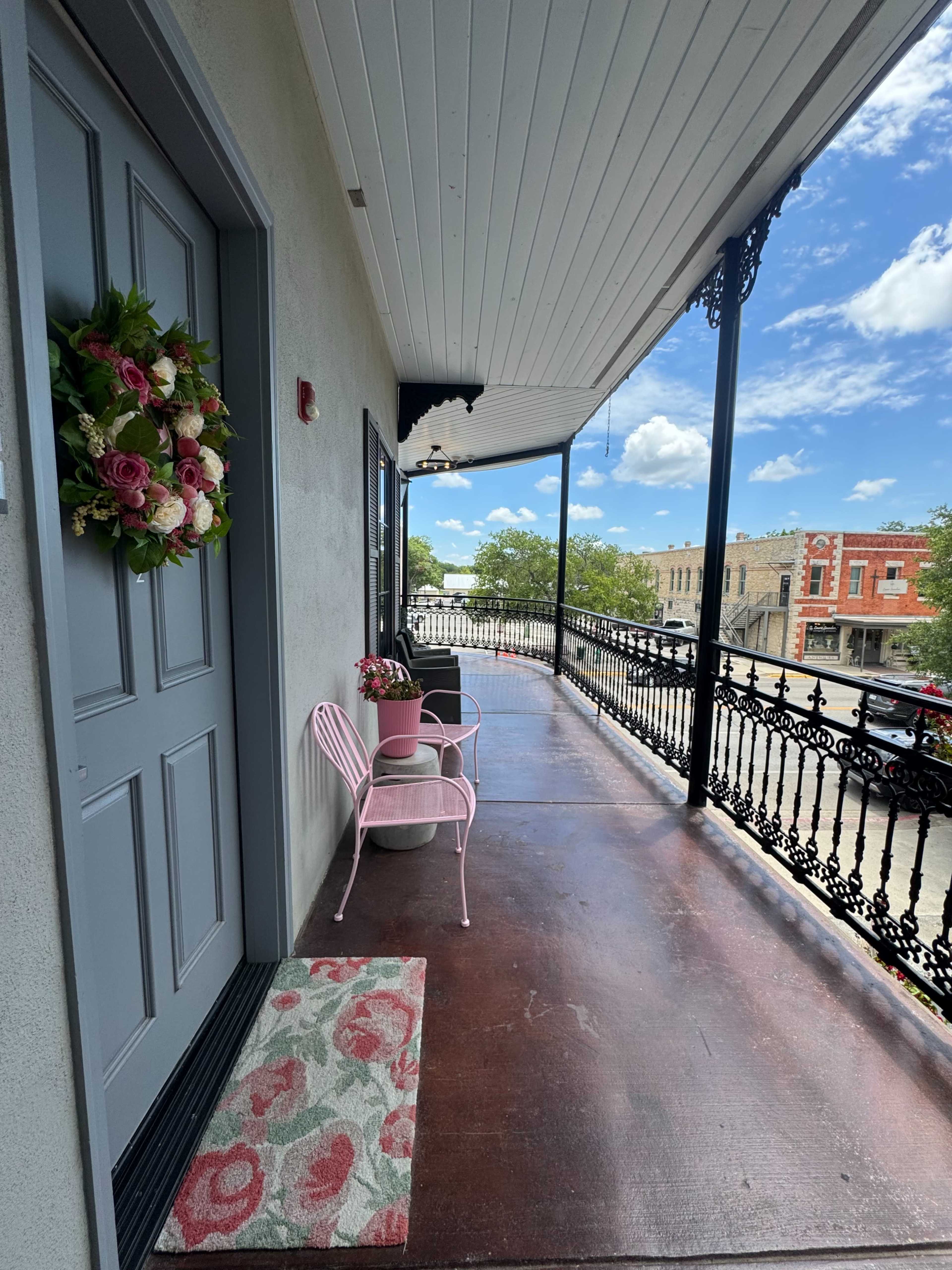 The image shows a charming balcony with a gray door, two pink chairs, and flower pots, overlooking a street lined with buildings under a partly cloudy sky.