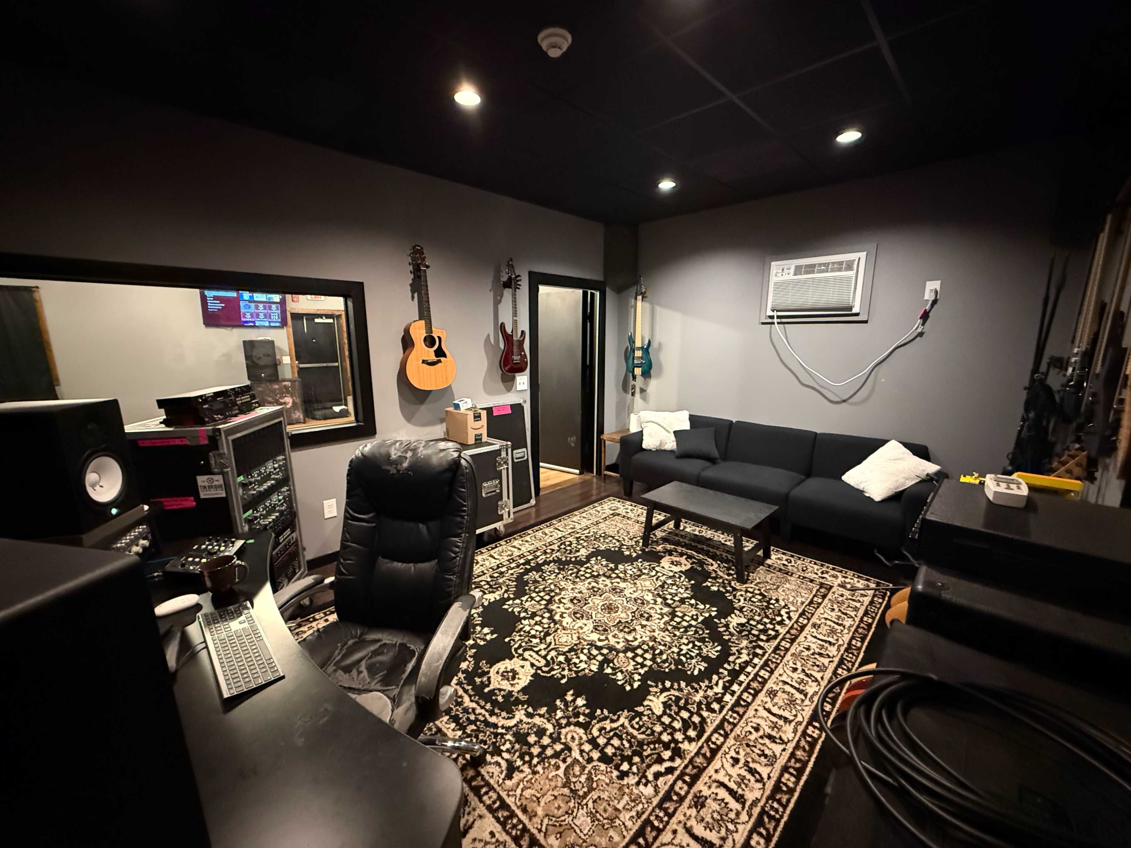 The image shows a music studio featuring a black couch, a patterned rug, acoustic instruments on the walls, and audio equipment.