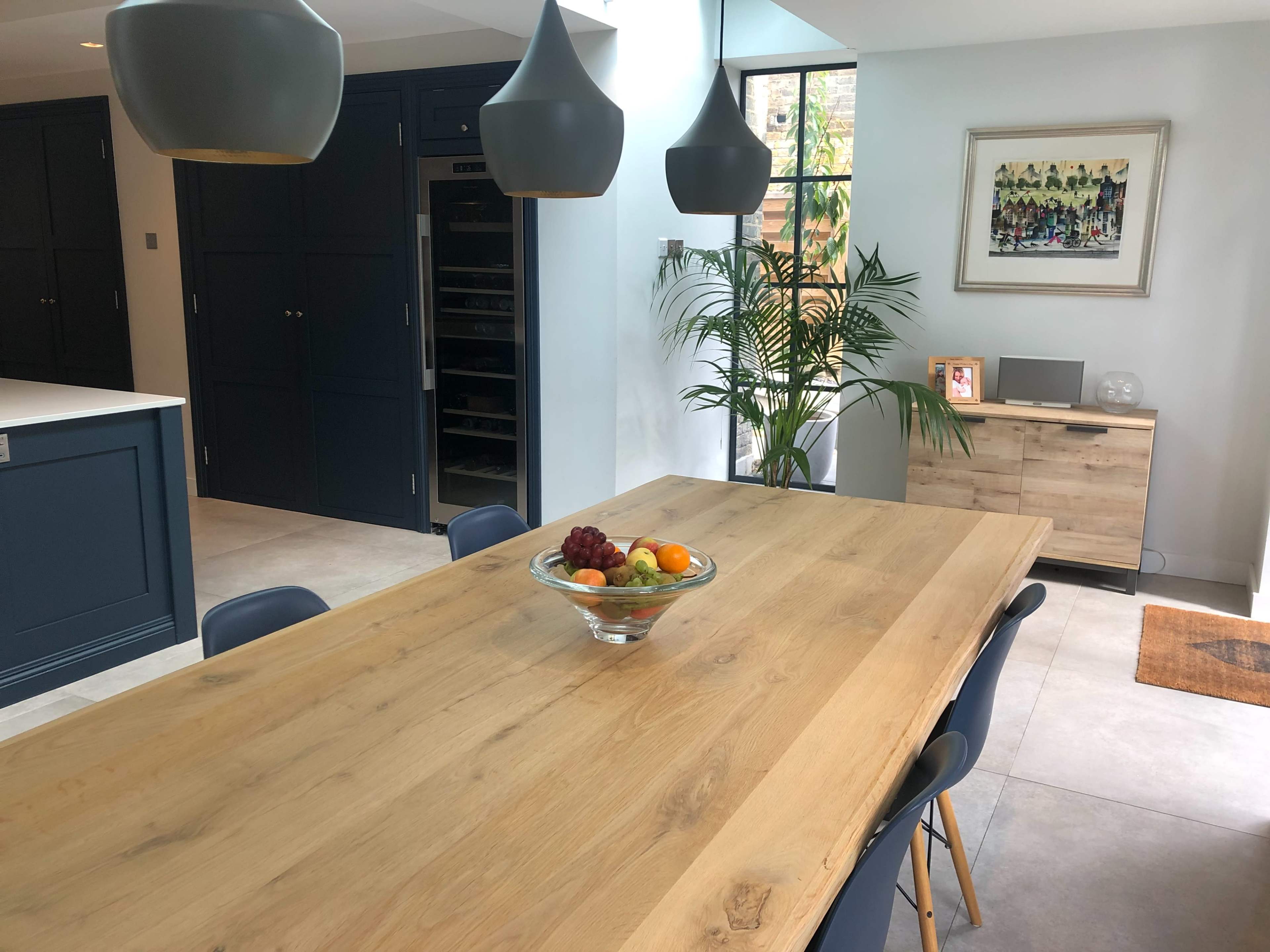 A dining table made of wood is centrally placed in a modern kitchen, surrounded by blue chairs, with a bowl of fruit on the table and a large window letting in natural light.