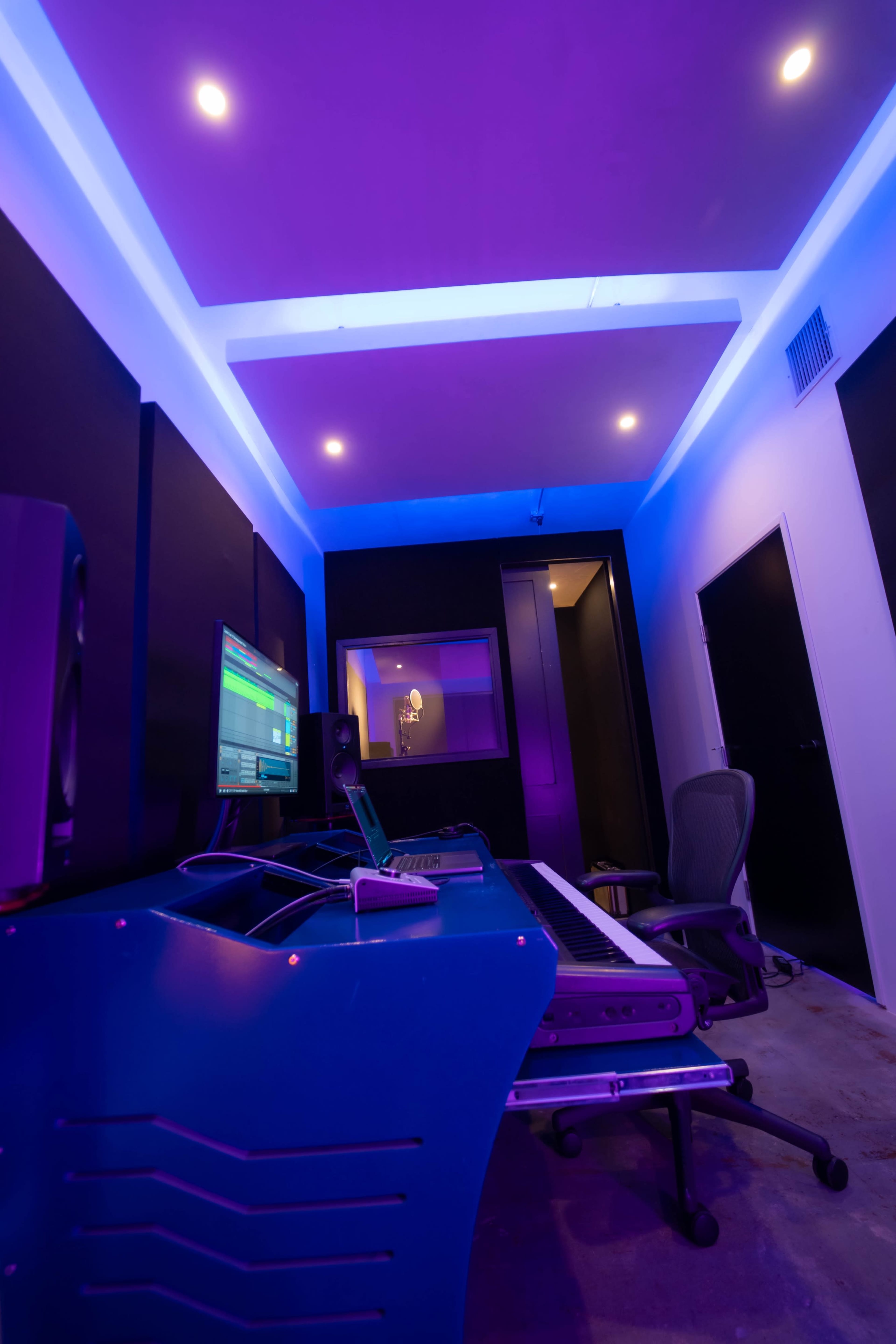 The image shows a modern music production studio with purple lighting, featuring a keyboard, computer setup, and a soundproof booth in the background.