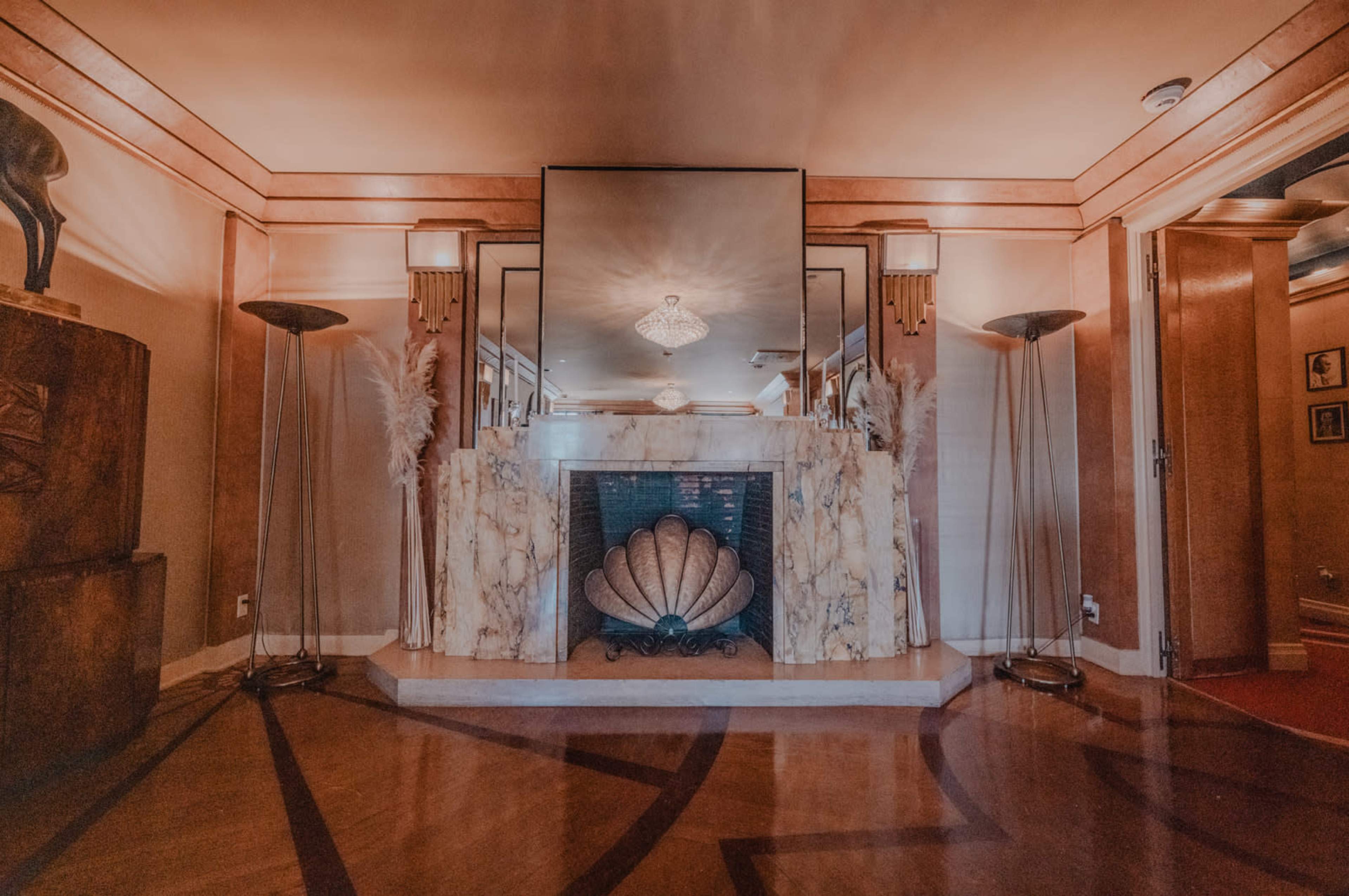 The image shows an art deco-style interior with a marble fireplace, a large mirror, and decorative lighting fixtures.