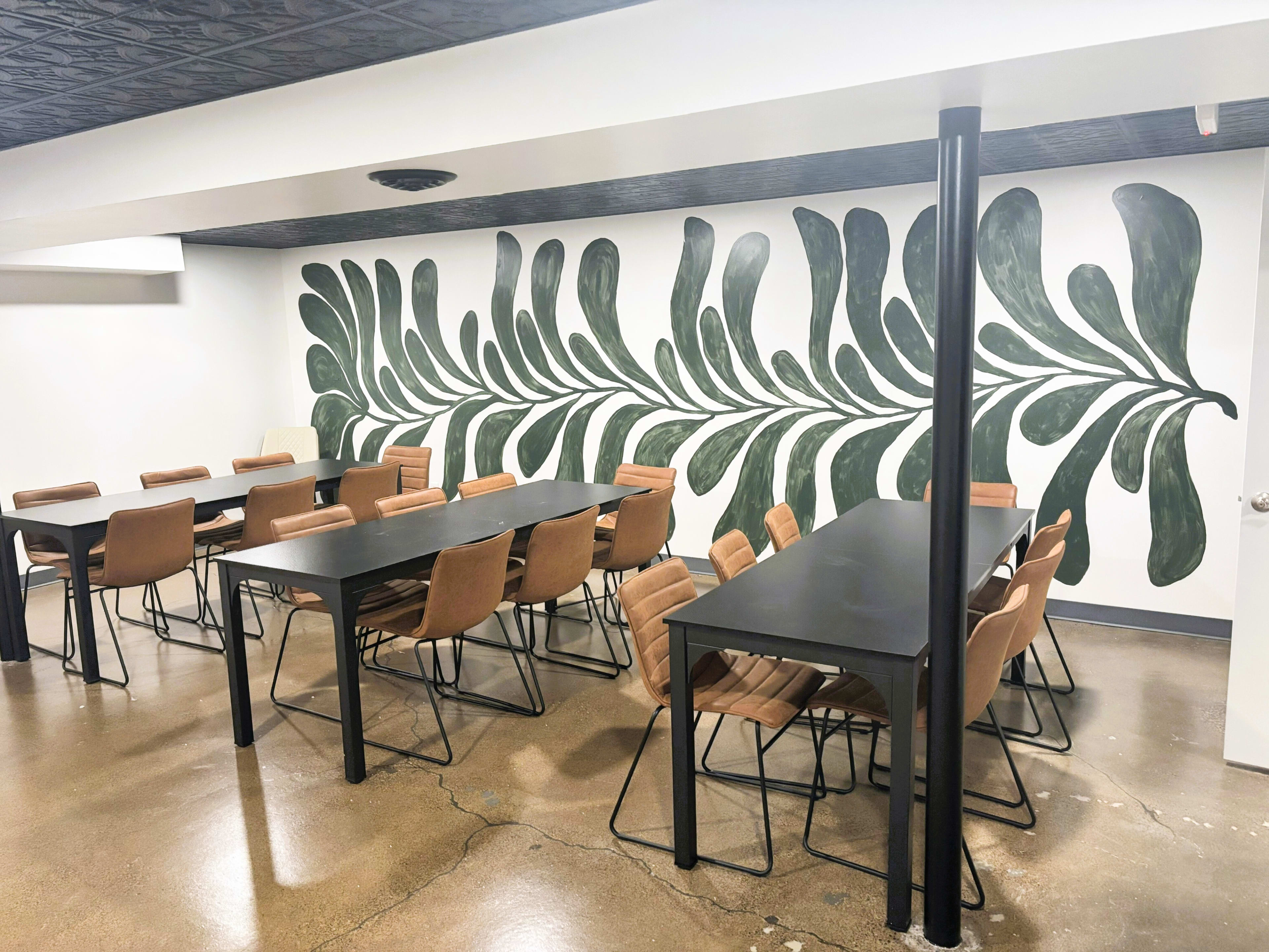 The image shows a modern conference room with a large mural of green leaves on one wall, featuring several rows of dark tables and brown chairs arranged neatly.