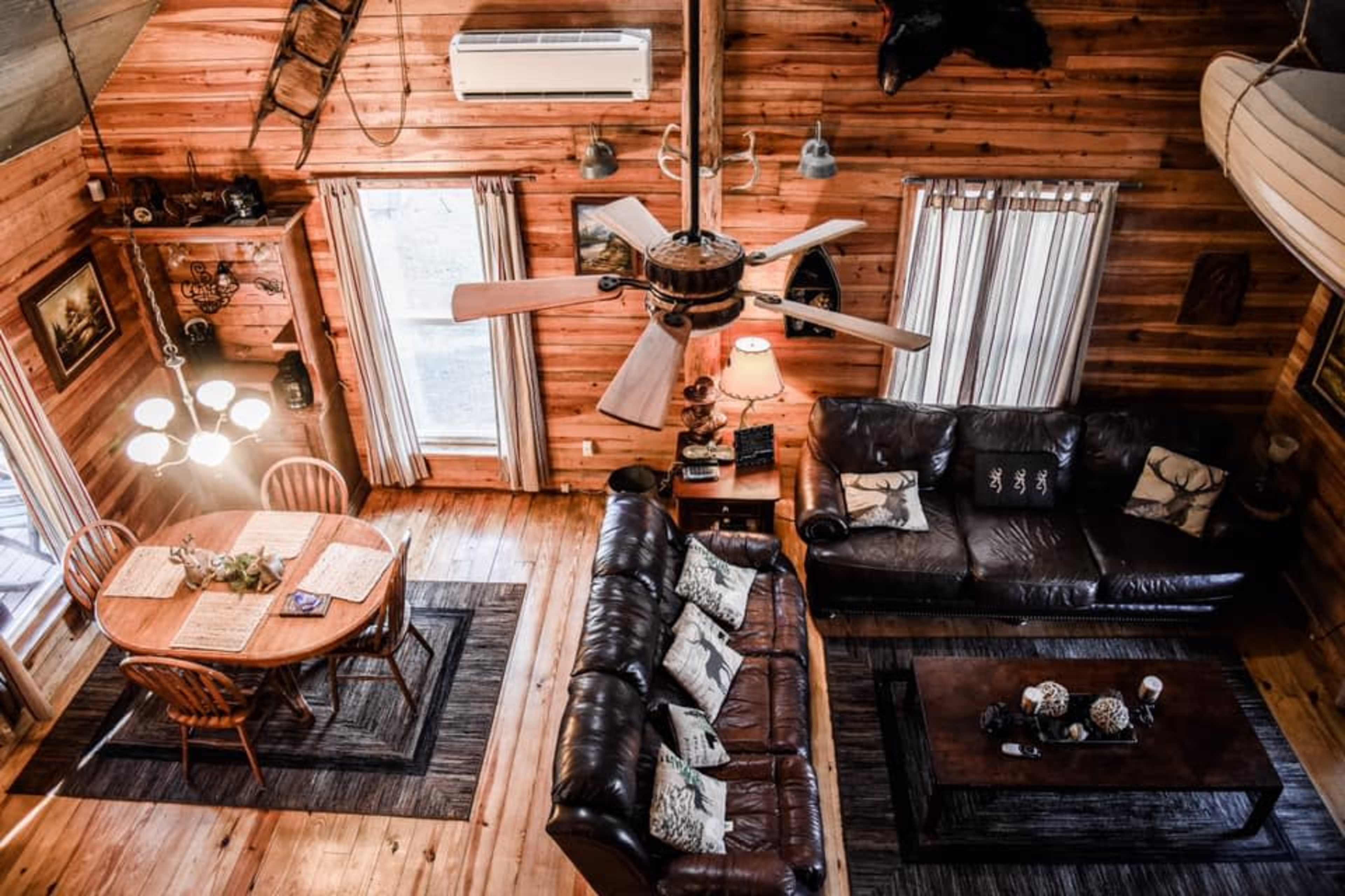 The image shows a rustic wooden cabin interior featuring a living area with two leather sofas, a wooden dining table, and ceiling fans.
