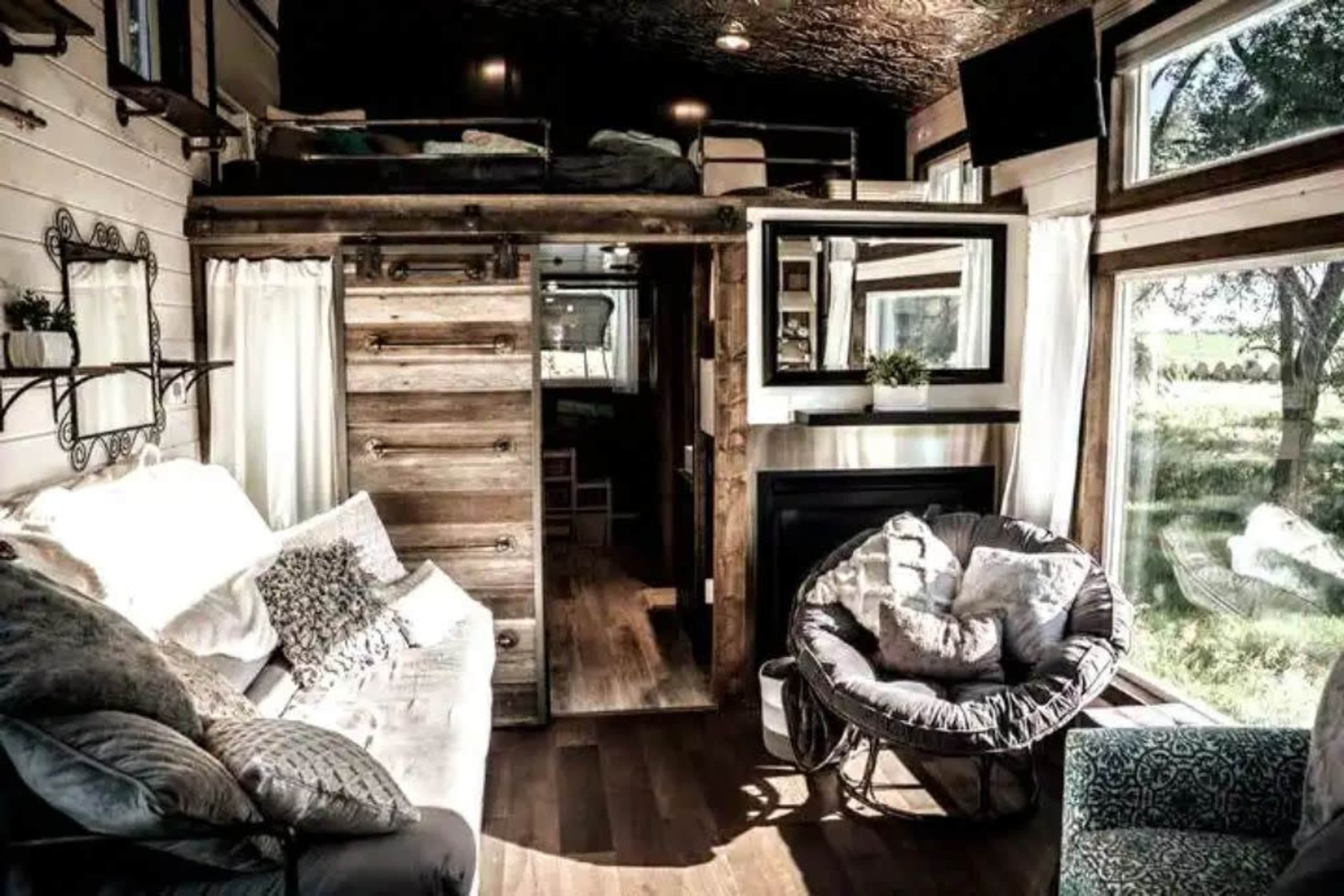 A cozy, modern tiny home interior with a lofted sleeping area, wooden accents, large windows, and a comfortable seating area.