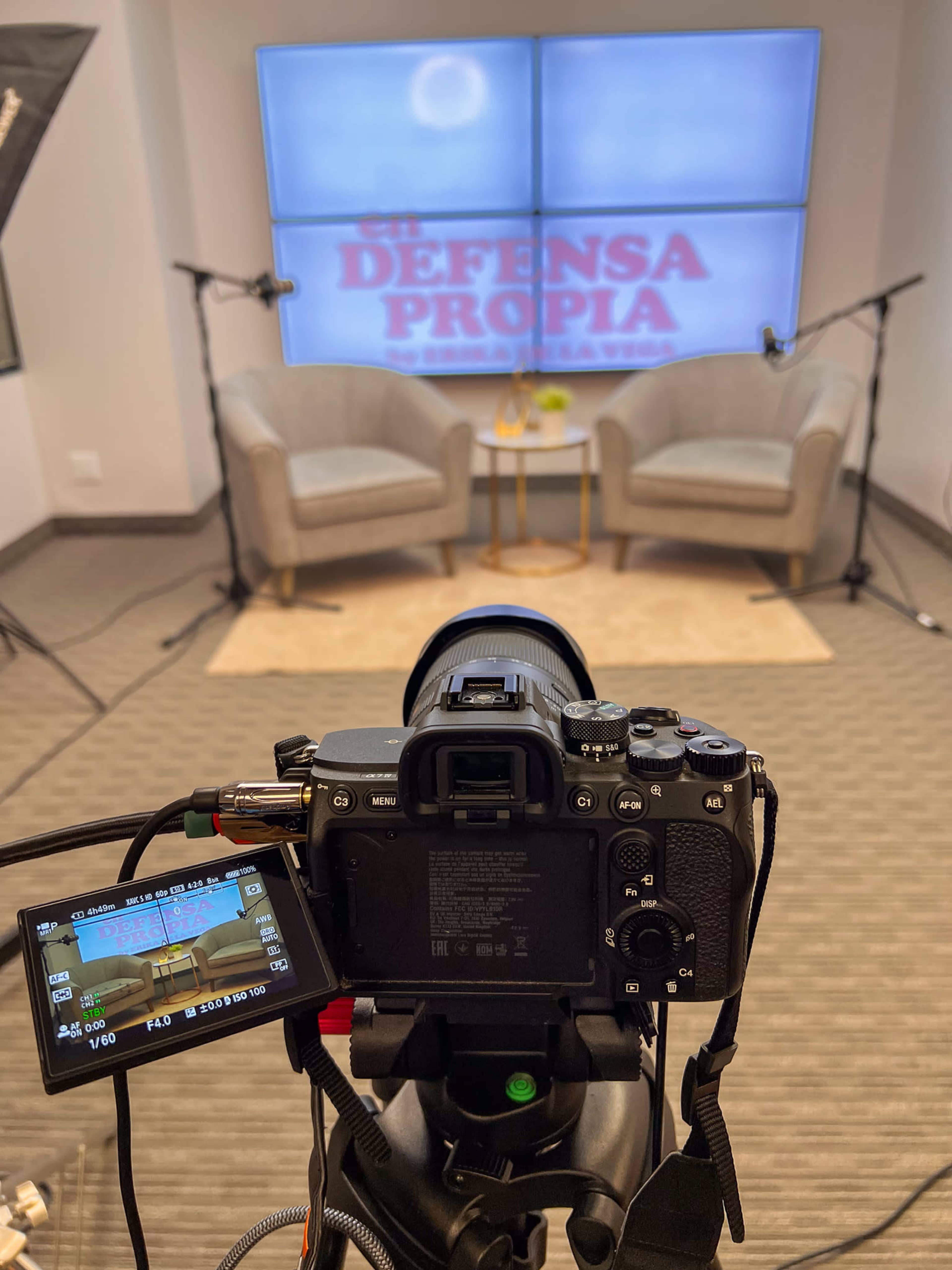 A camera is set up on a tripod facing a staged interview setup with two chairs and a large screen displaying the words "El Defensa Propia."