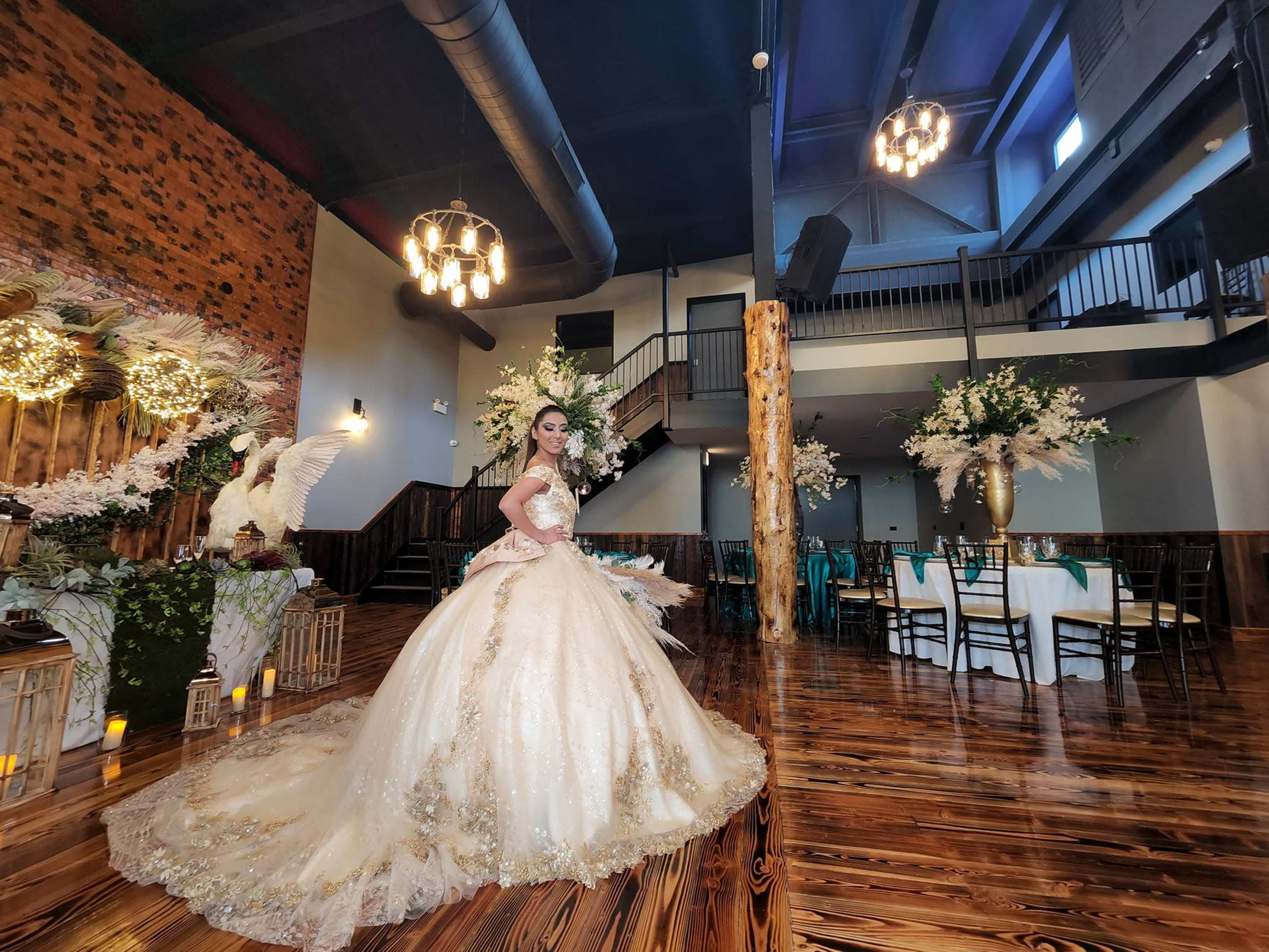 A bride in a golden wedding gown poses in a decorated reception venue with floral arrangements and elegant lighting.