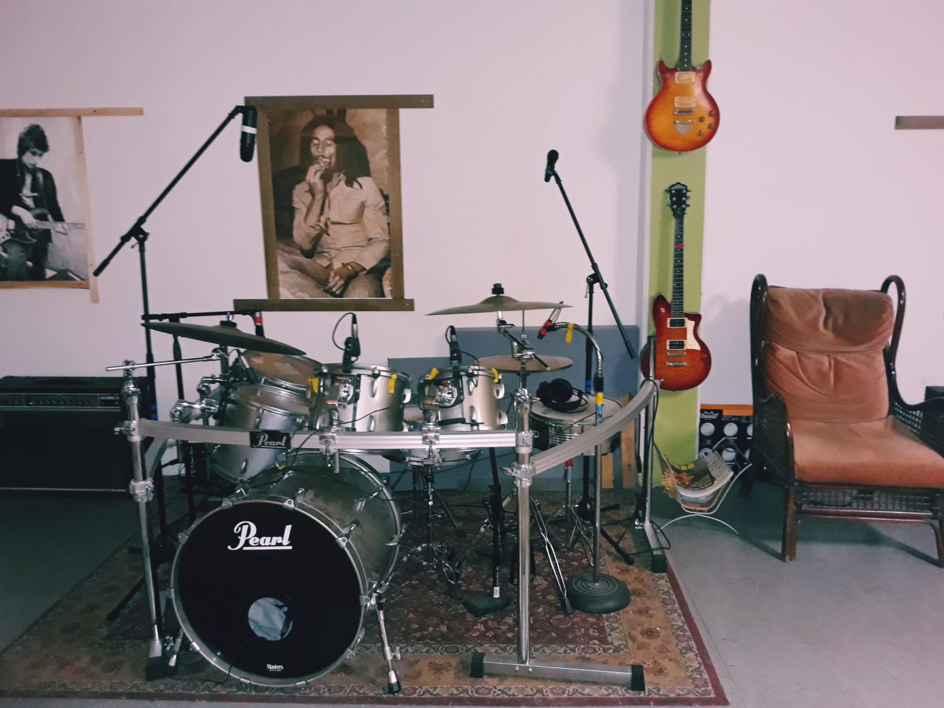A drum set is arranged in a room with hanging guitars, a microphone setup, and framed photographs on the walls.