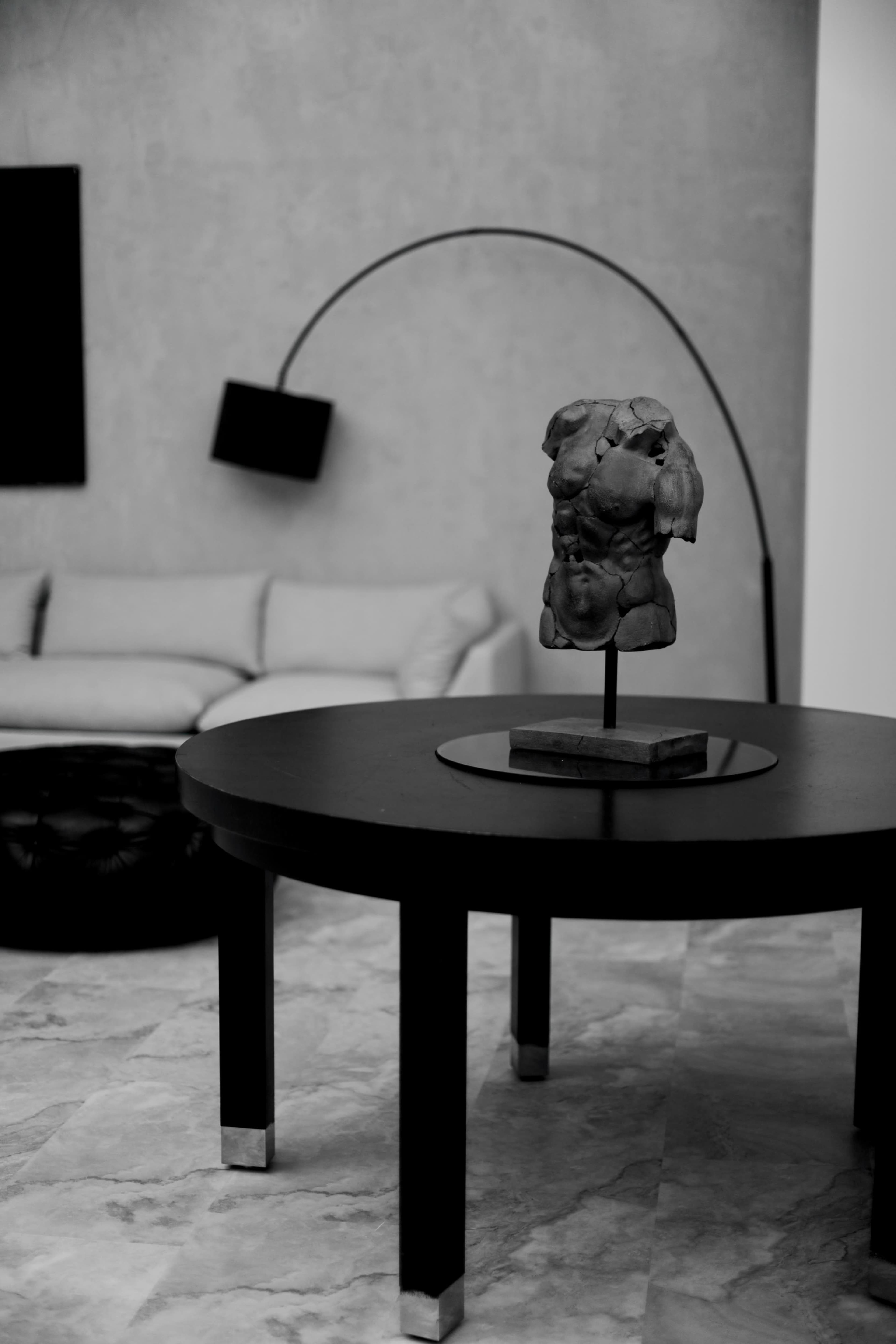A sculptural torso stands on a circular black table in a minimalist room with a sofa and a floor lamp in the background.