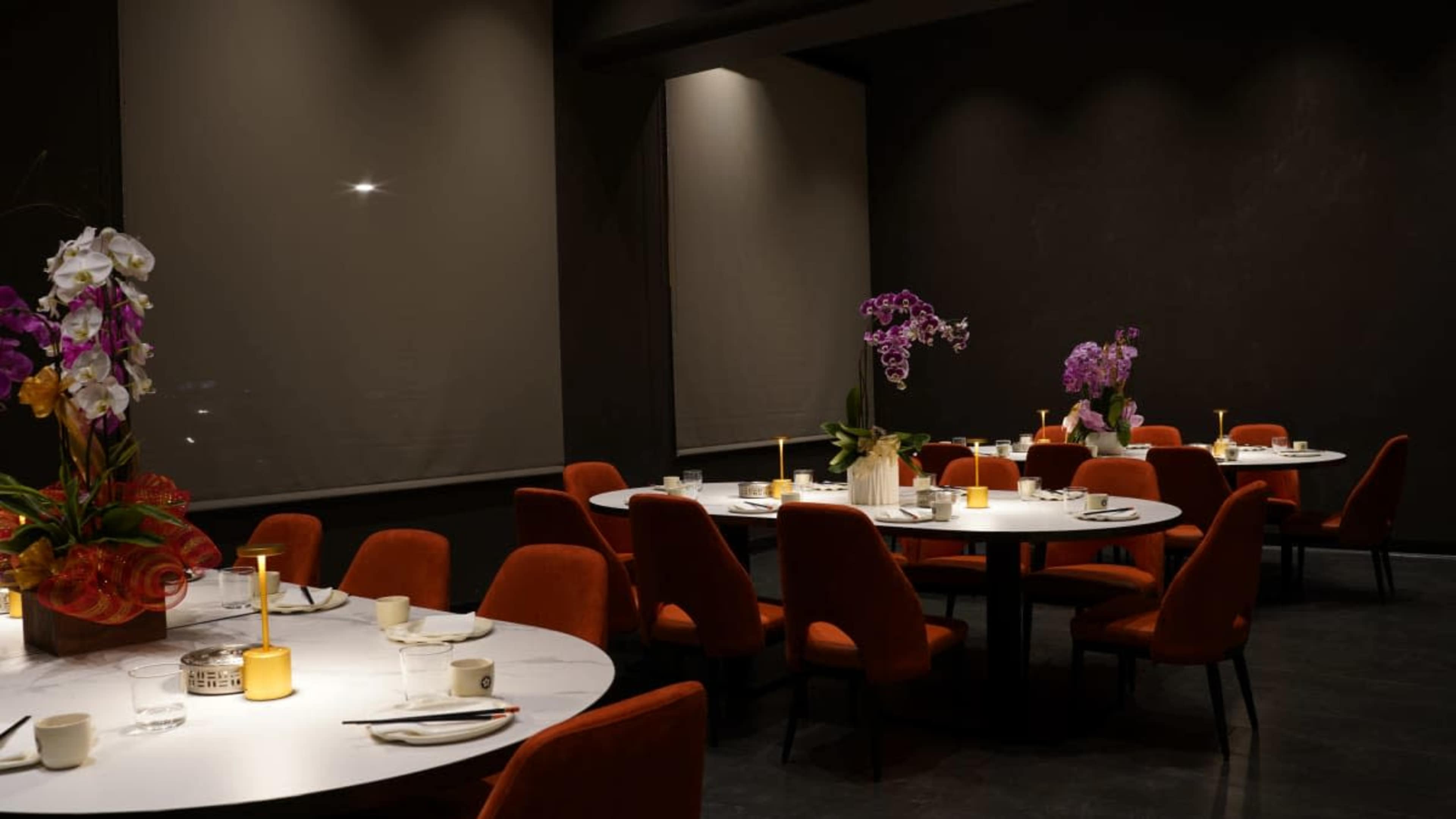 A modern restaurant interior features several round tables set for dining, adorned with floral centerpieces and illuminated by soft candlelight.