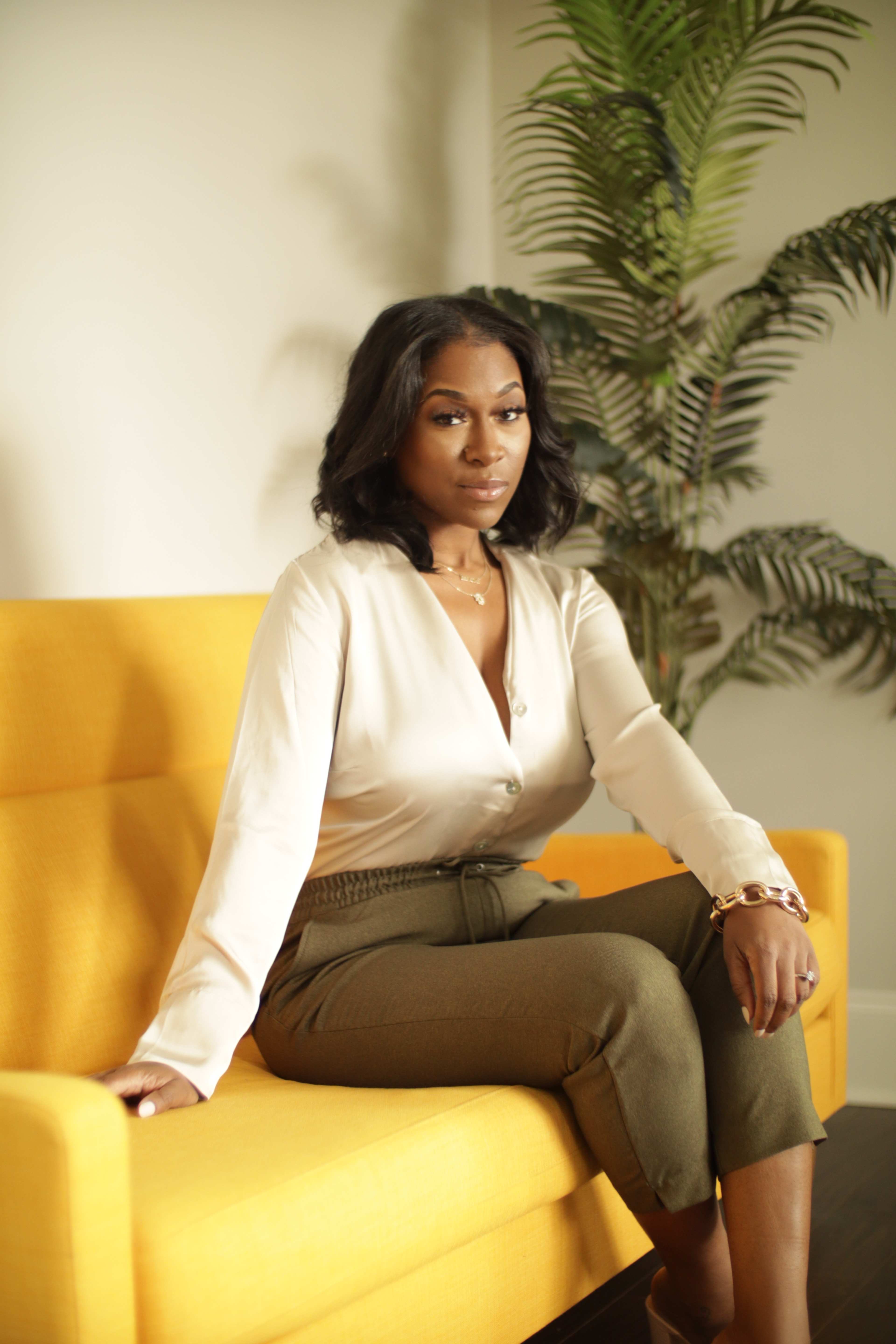 A woman in a beige blouse and olive green pants sits on a yellow sofa in a well-lit room with a potted plant.