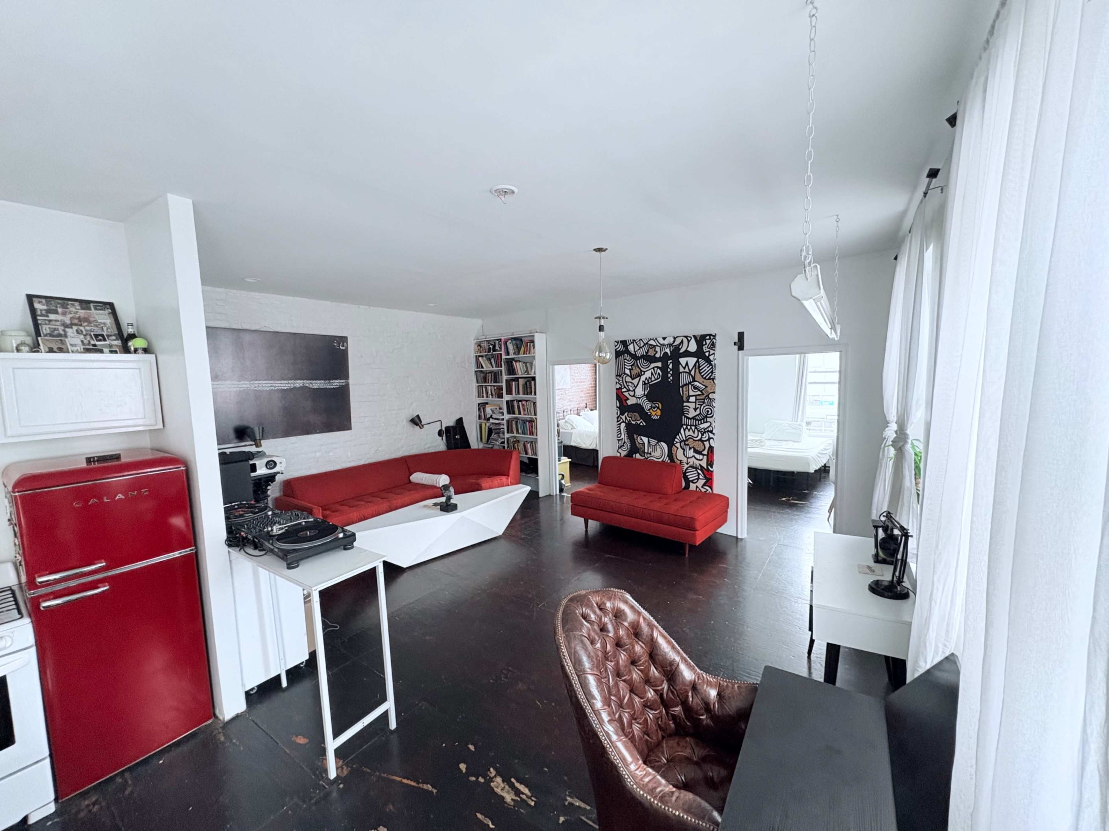 The image shows a spacious, modern living area featuring a red kitchen appliance, a white wall with bookshelves, and various seating areas including red couches.