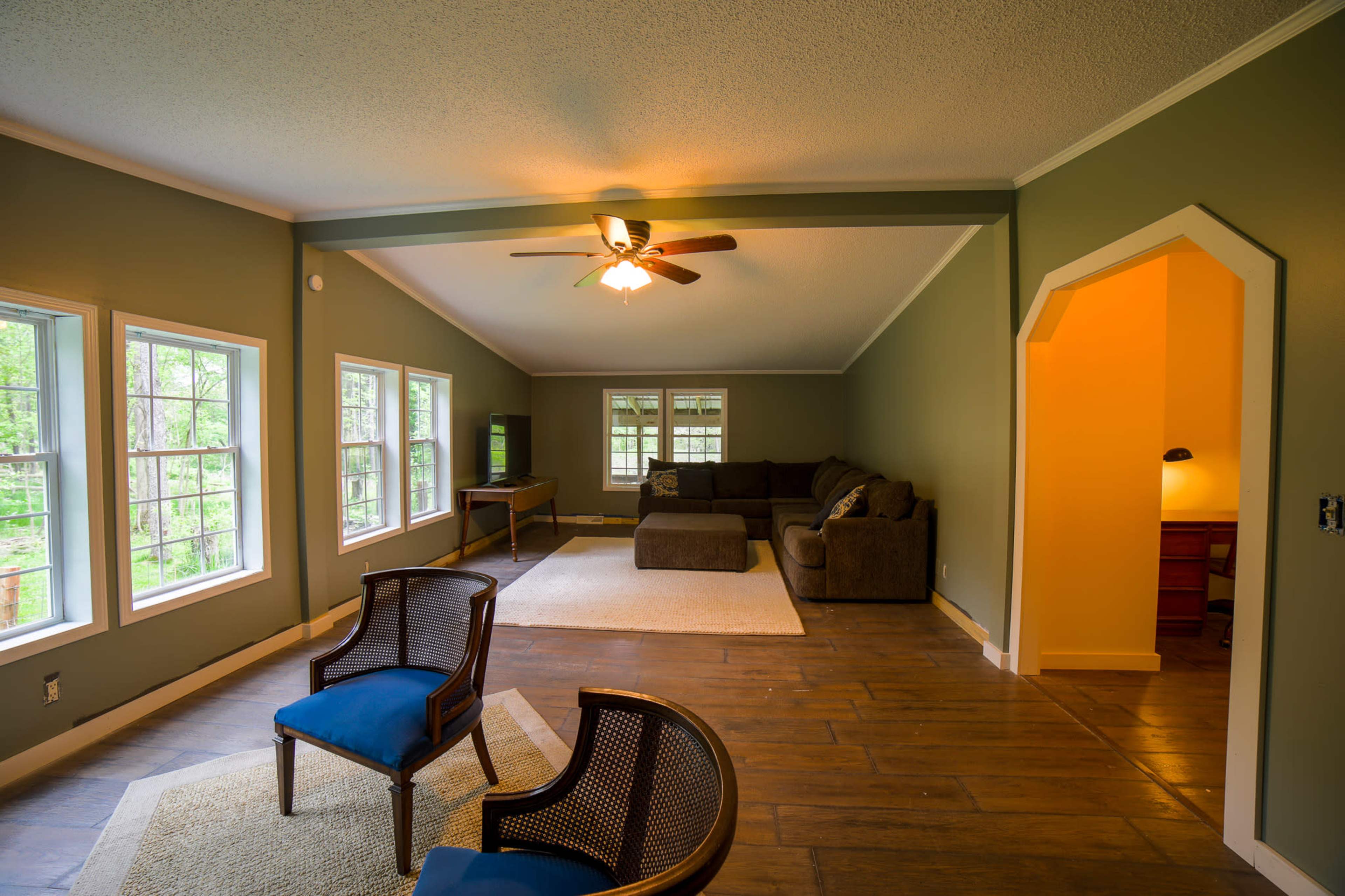 The room features a spacious, open layout with large windows, a ceiling fan, and a mix of seating areas including a couch and two chairs.