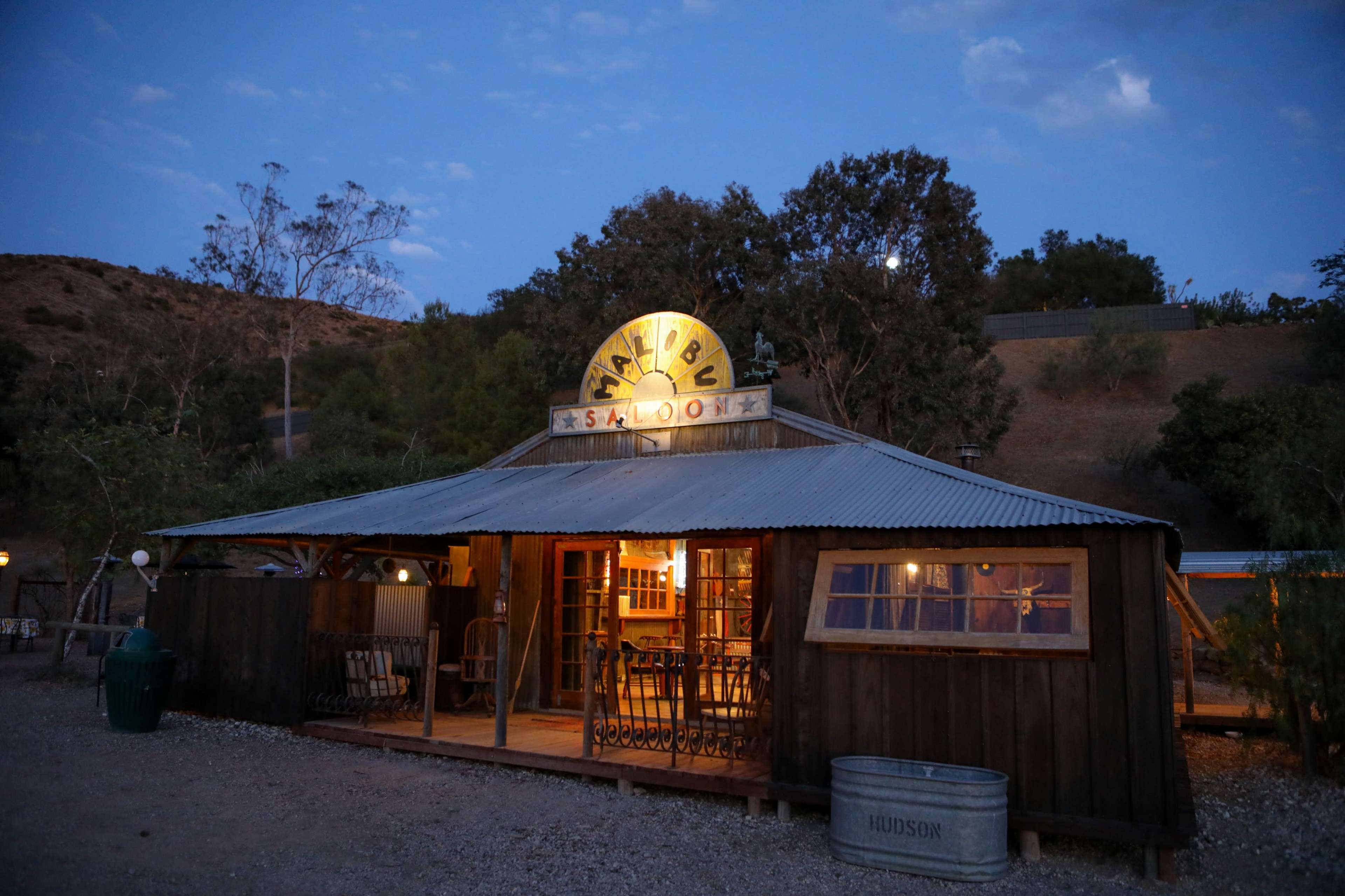 A rustic wooden saloon with a corrugated metal roof stands in a dusk setting, illuminated by a round sign that reads "Malibu Saloon."