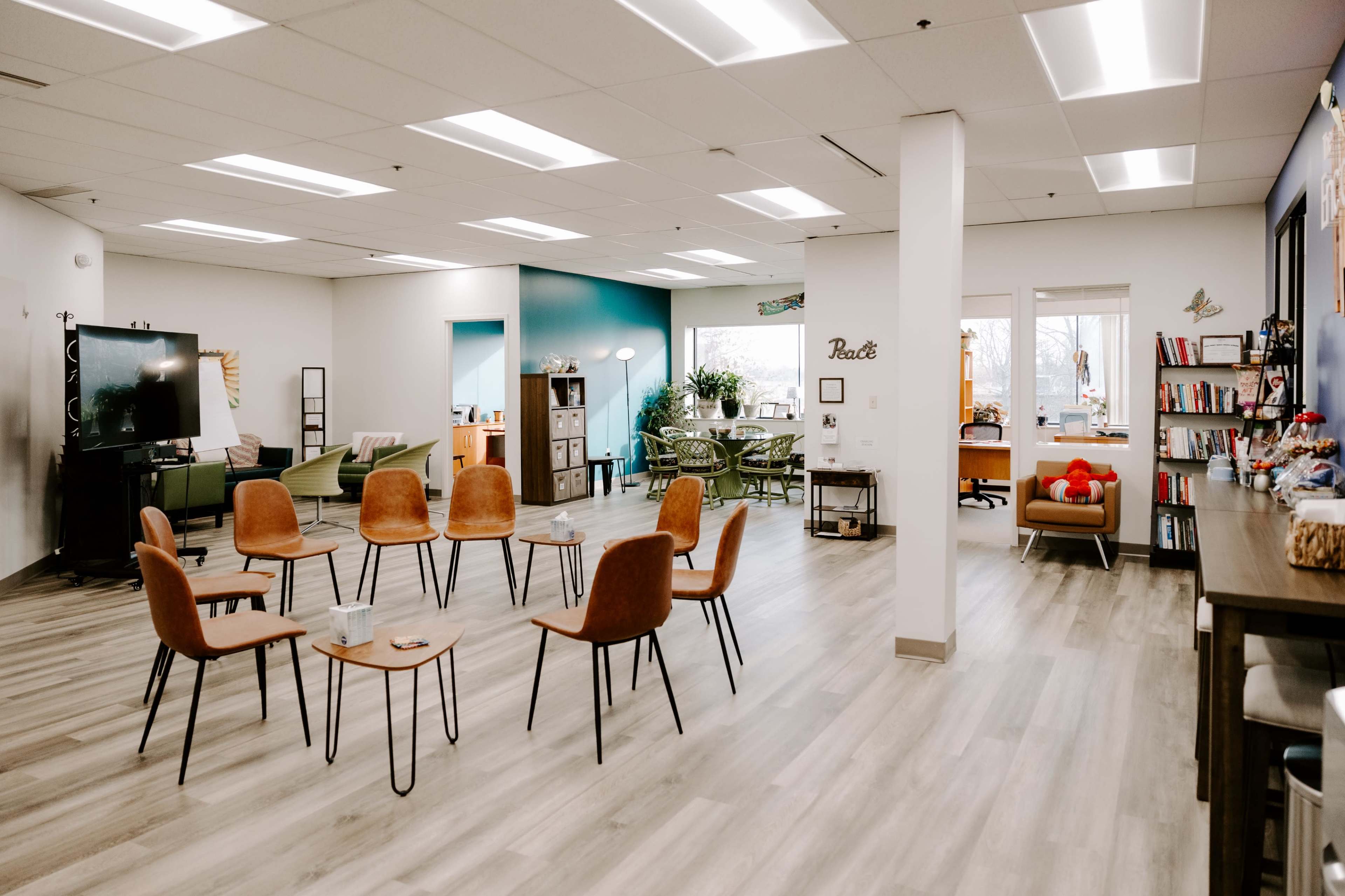 The image shows a spacious room with a circle of chairs around small tables, complemented by various seating areas and shelves filled with books.