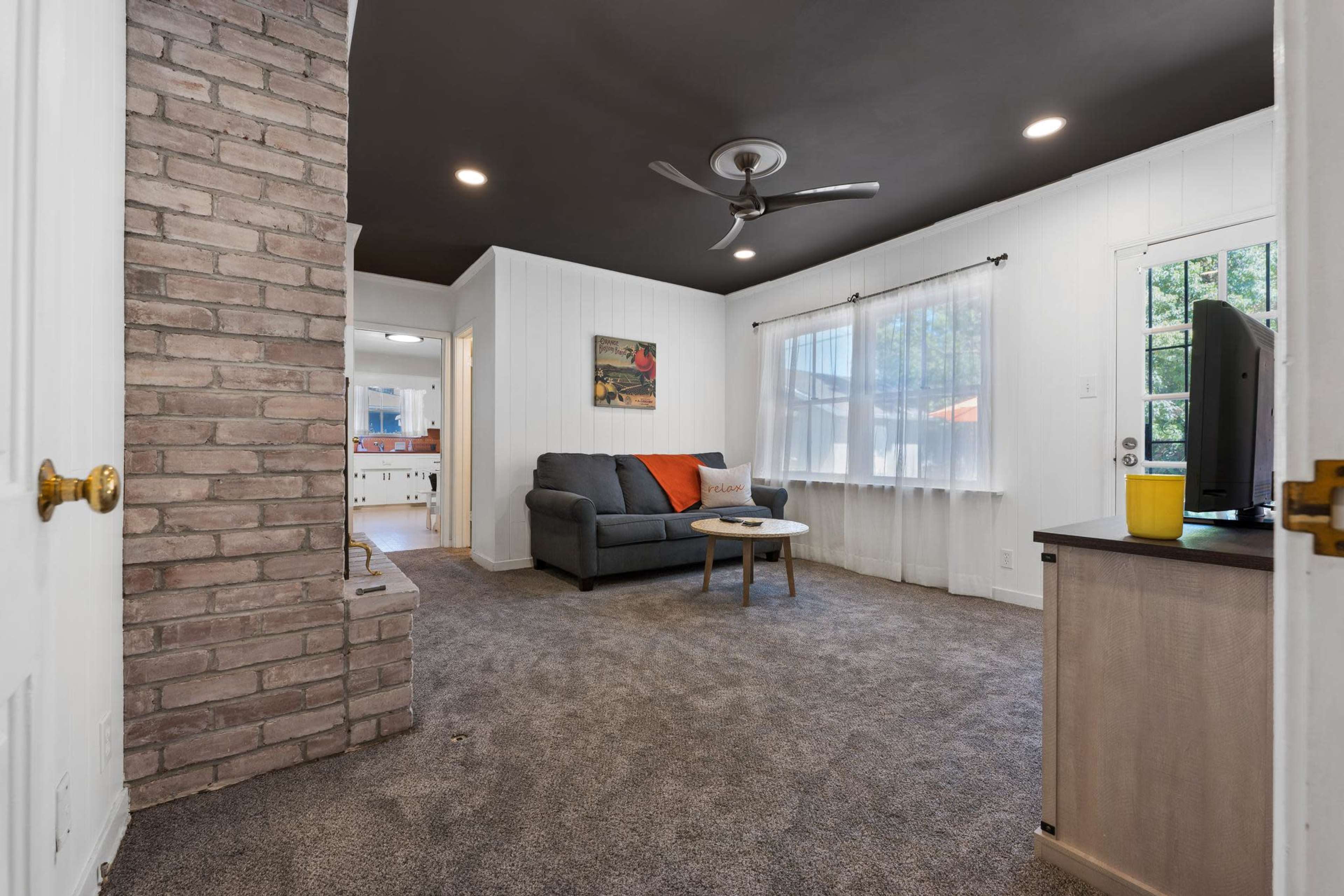 A living room with a gray couch and an orange accent pillow, a small coffee table, and a brick fireplace, all under a dark ceiling.