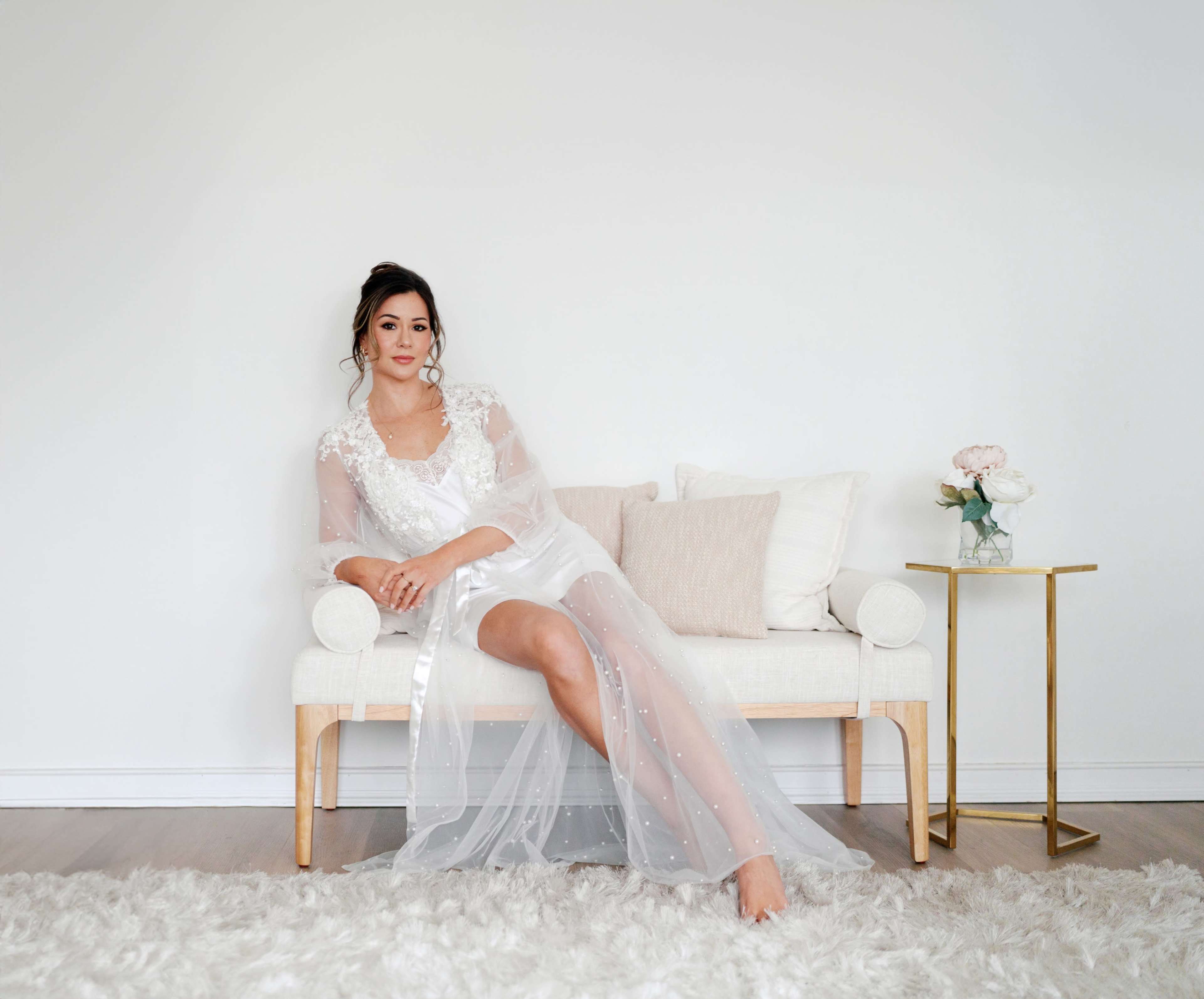 A woman in a white gown sits gracefully on a light-colored sofa with a soft rug beneath her.