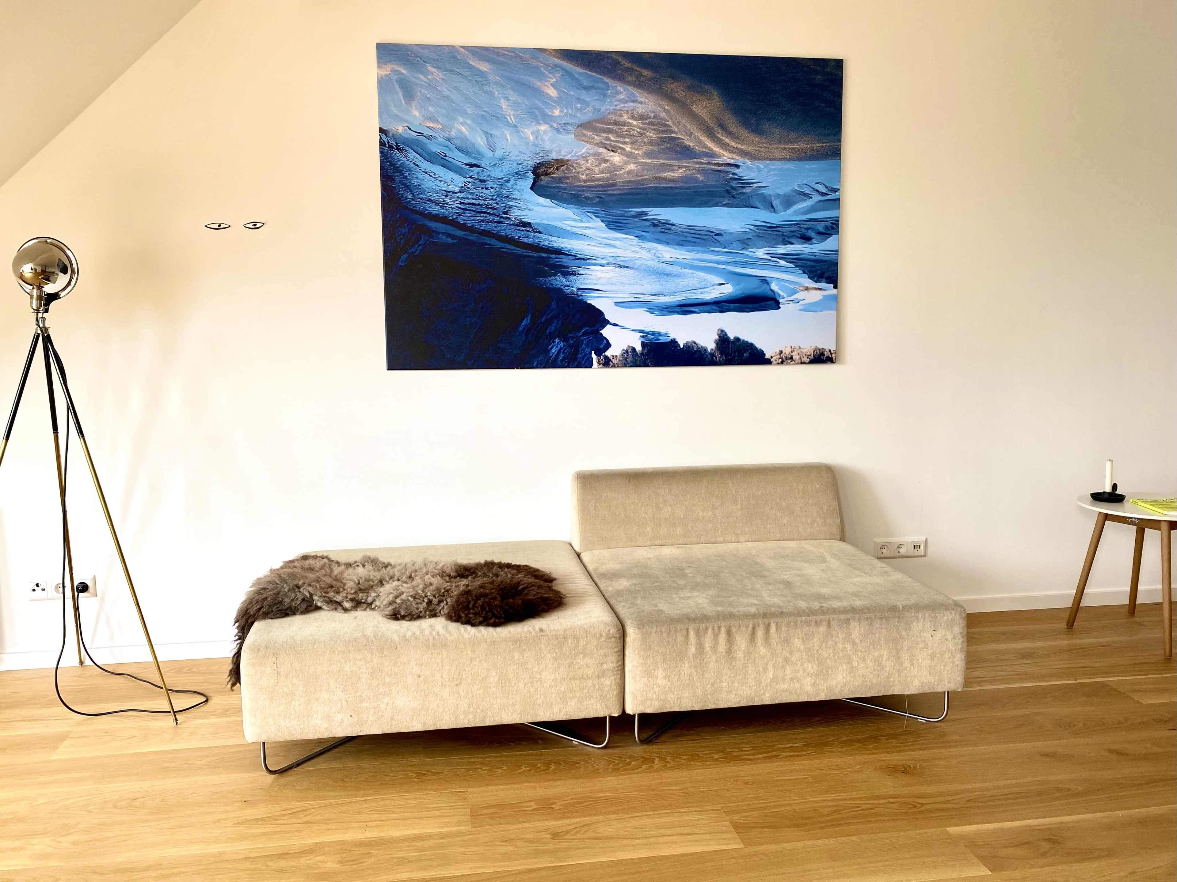 A minimalist living space featuring a beige sectional sofa, a floor lamp, a wooden side table, and a large landscape painting on the wall.