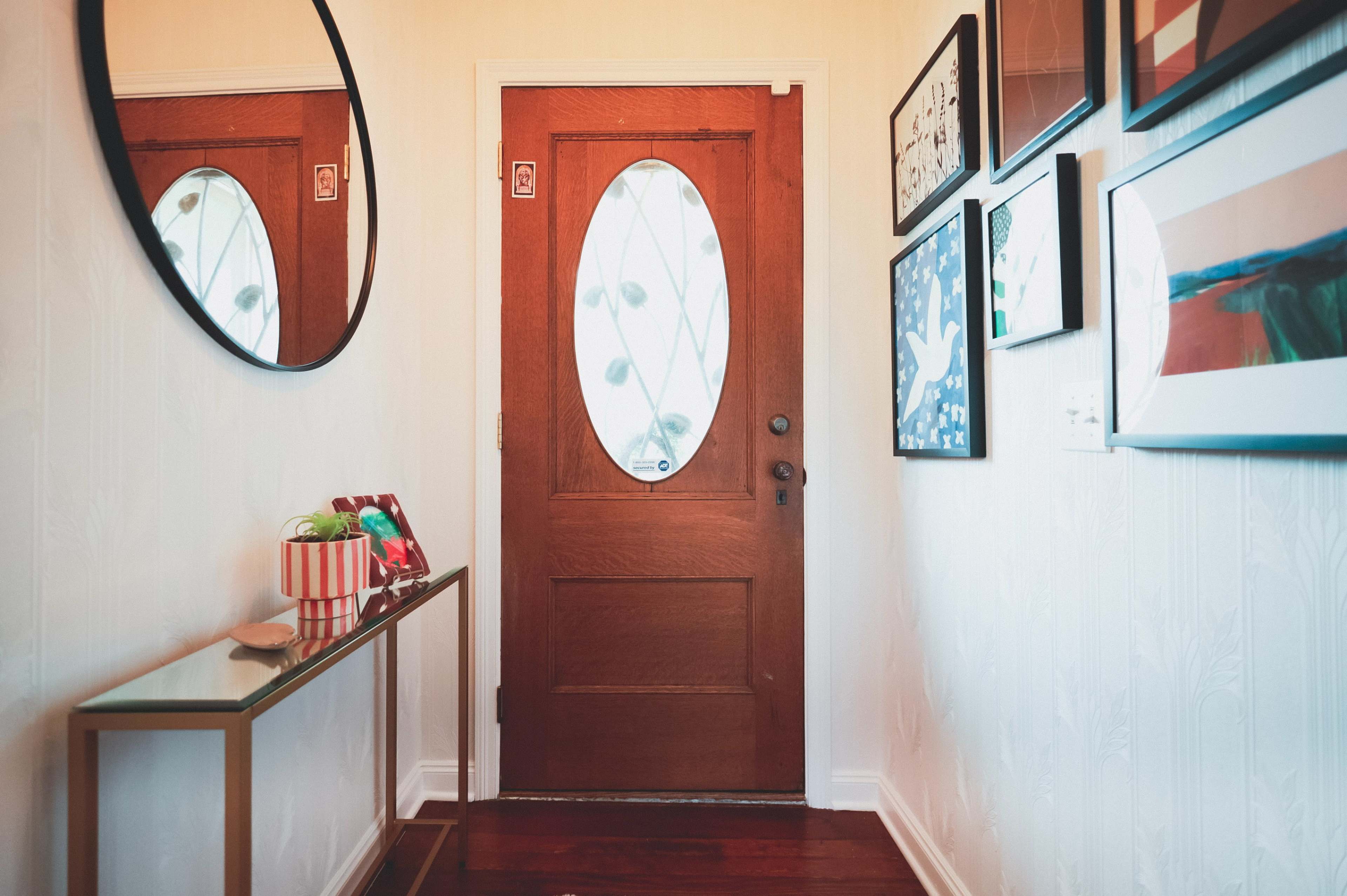 A wooden door with an oval window is positioned at the end of a narrow hallway, adorned with framed art on the walls and a glass console table on one side.