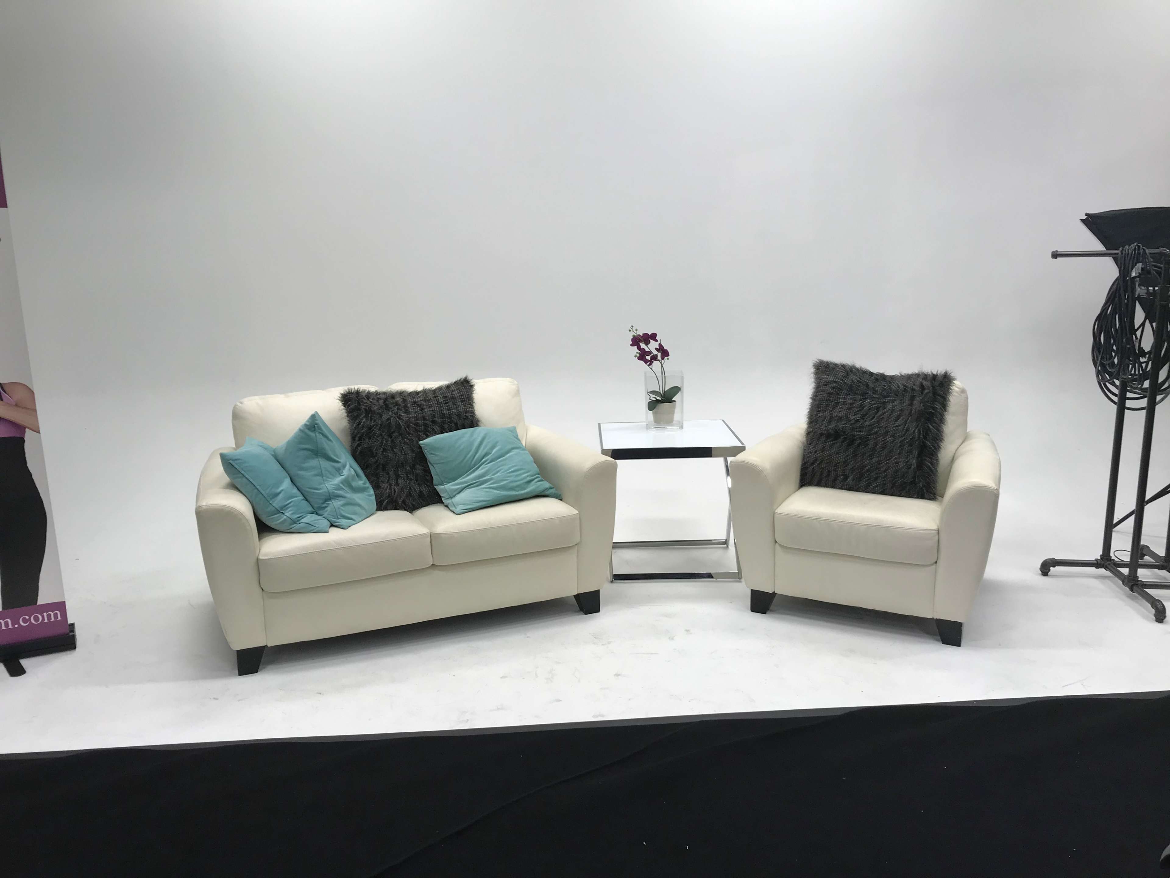 A white couch and a matching armchair with turquoise cushions are positioned beside a glass side table and a small potted orchid in front of a plain white backdrop.