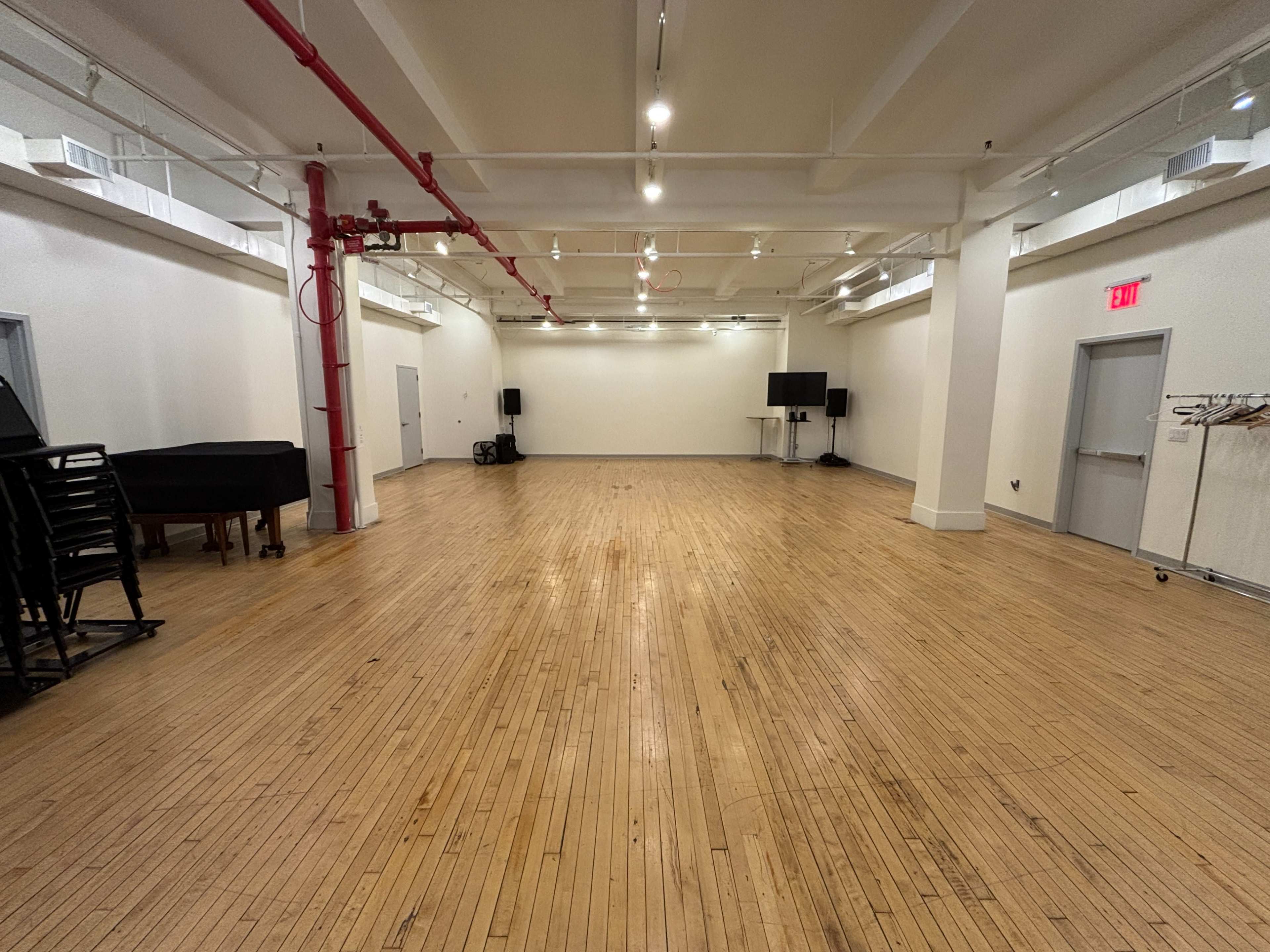 The image shows a large, empty room with wooden floors and white walls, featuring a red pipe along one side and equipment such as a piano and speakers in the background.