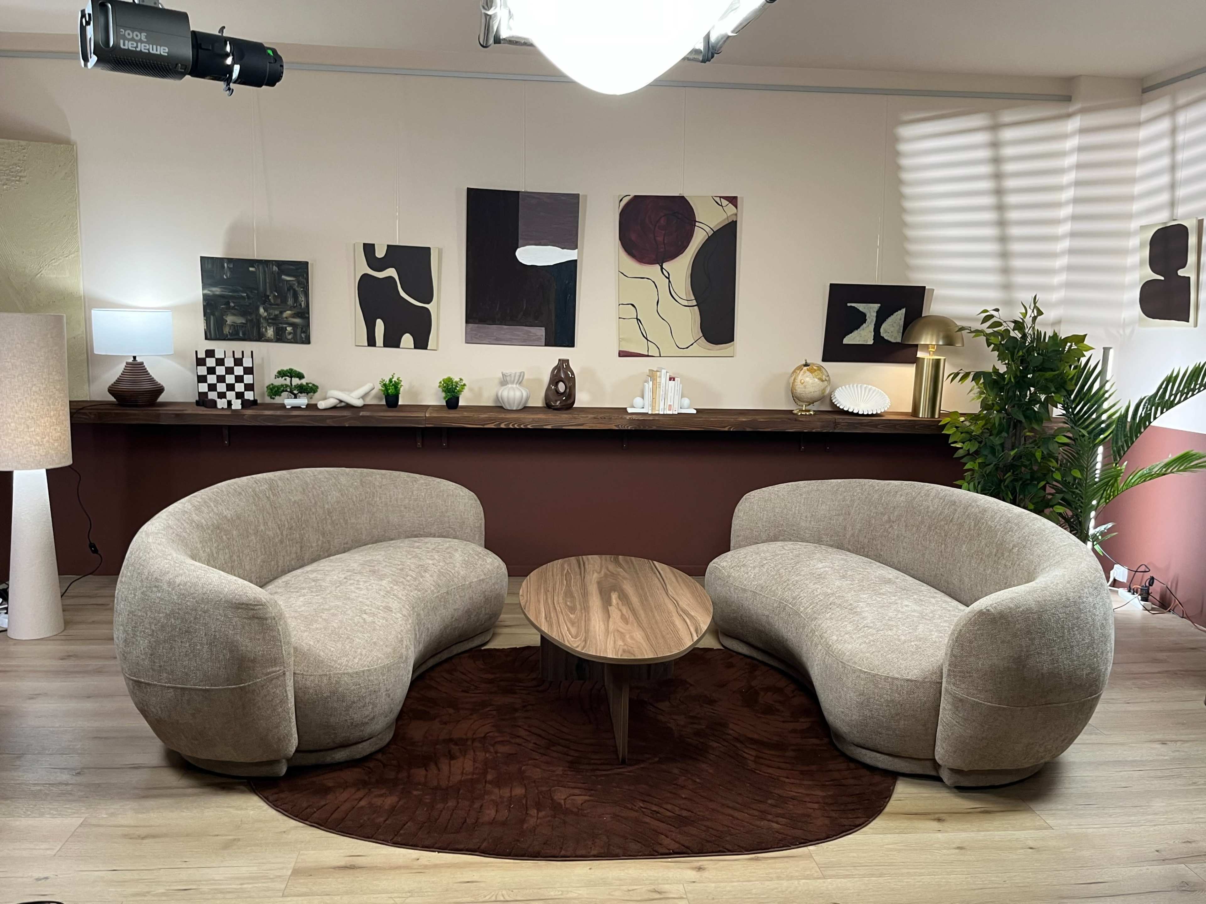 The image shows a modern interior space featuring two curved beige sofas positioned around a round wooden coffee table, with abstract artwork and decorative items on the walls and shelves.