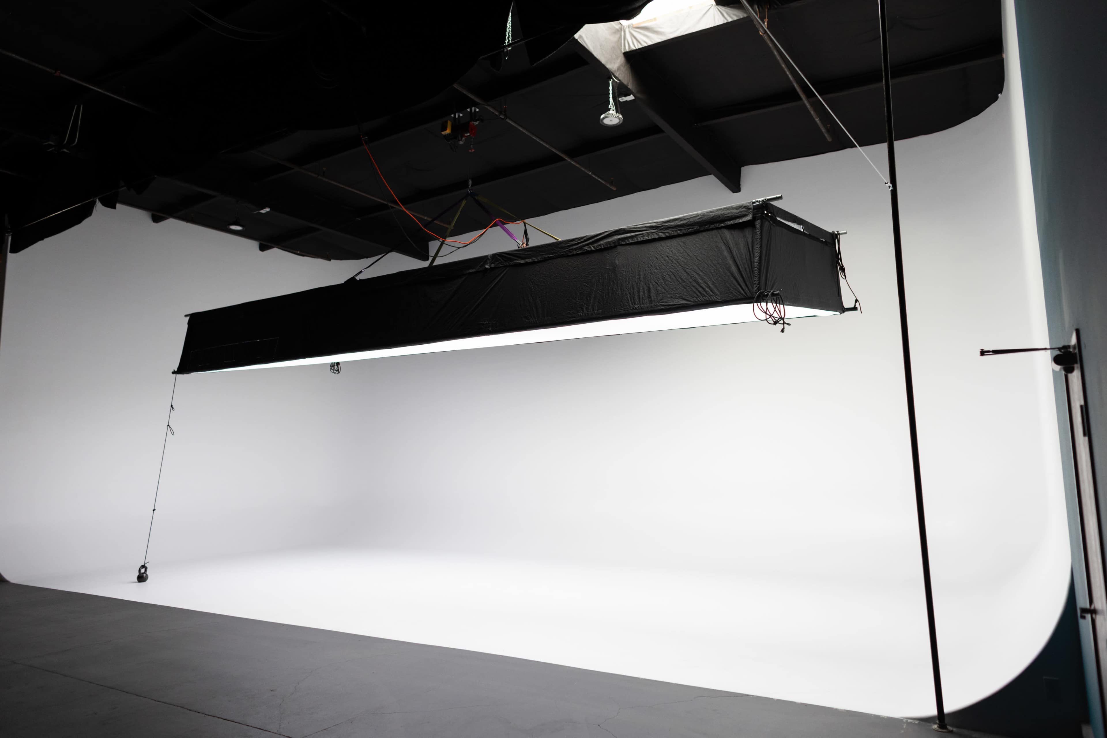The image shows an empty photography studio with a large softbox light suspended from the ceiling above a seamless white background.