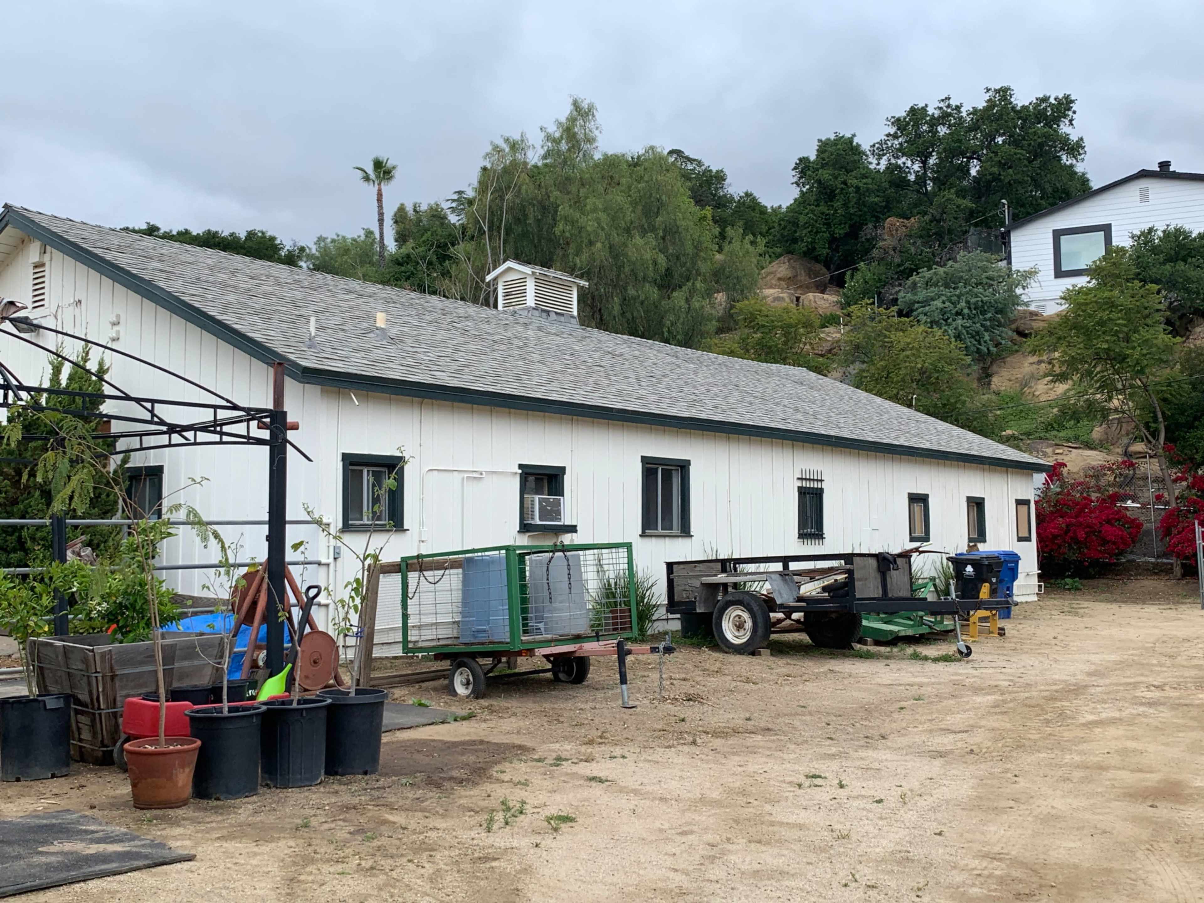 The image shows a single-story white building with a gray roof, surrounded by outdoor equipment and potted plants on a dirt lot.
