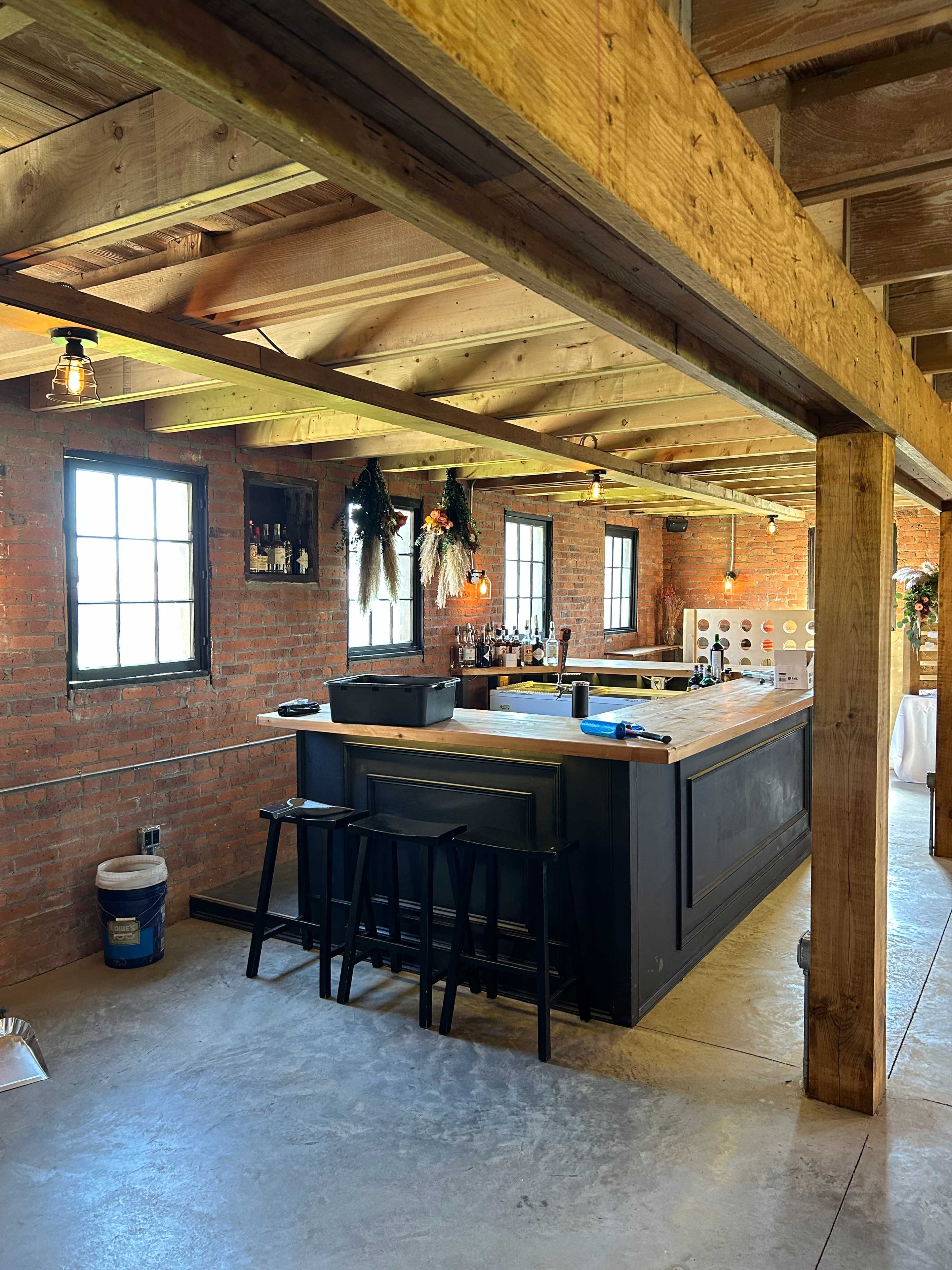 A rustic bar area with a wooden counter, black stools, and exposed brick walls beneath a wooden ceiling.
