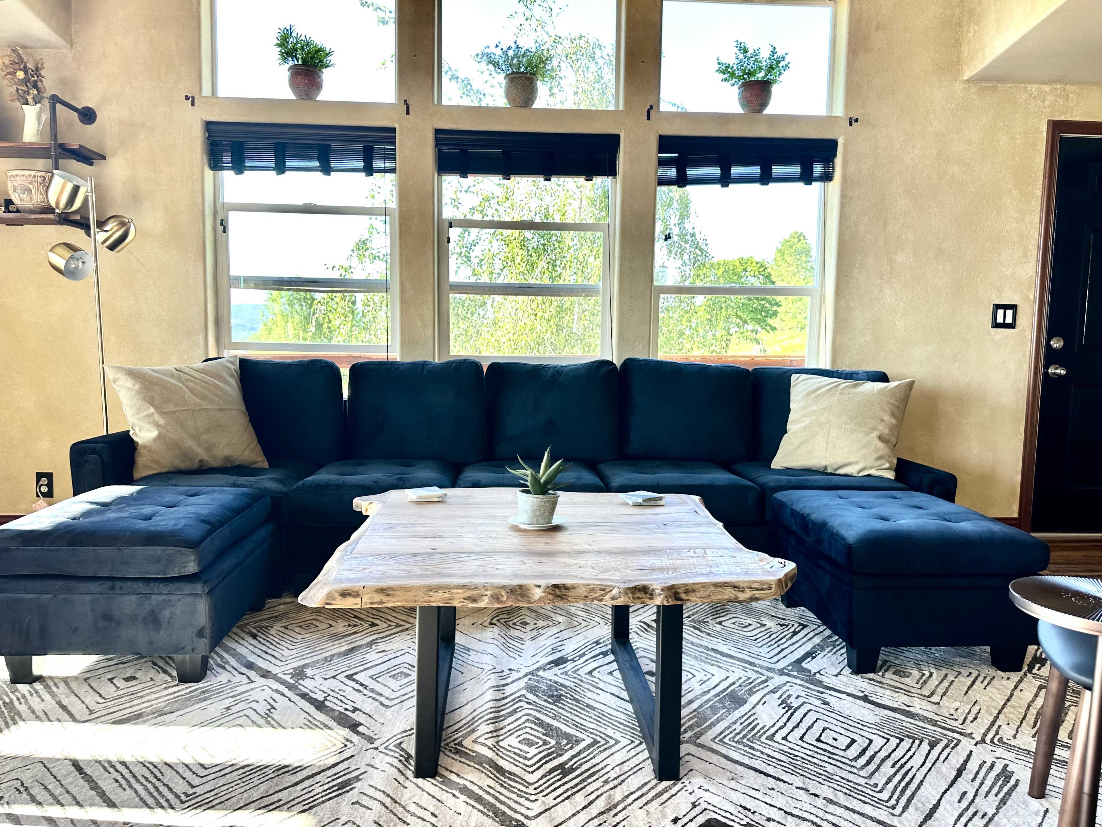 The image shows a bright living room featuring a dark sectional sofa with two pillows, a wooden coffee table with black legs, and large windows with potted plants on the ledges.