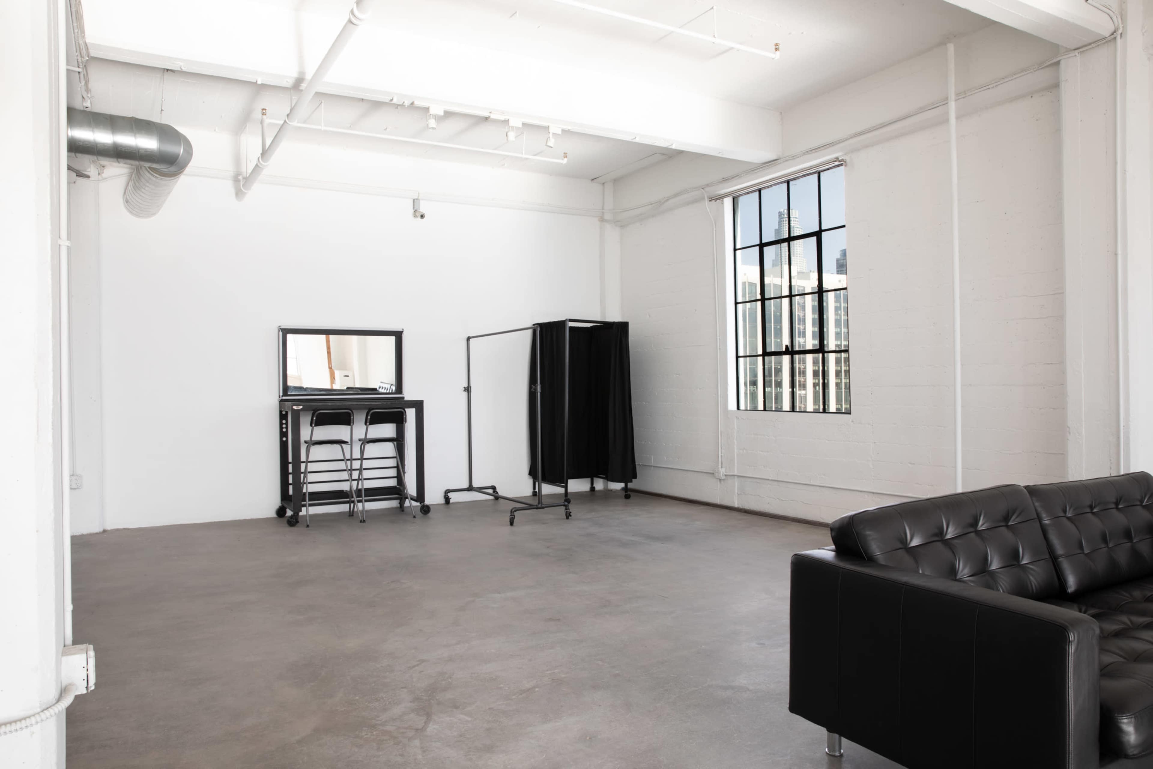 The image shows a spacious, empty studio with a large window, a black couch, a clothing rack, and a mirrored stand.