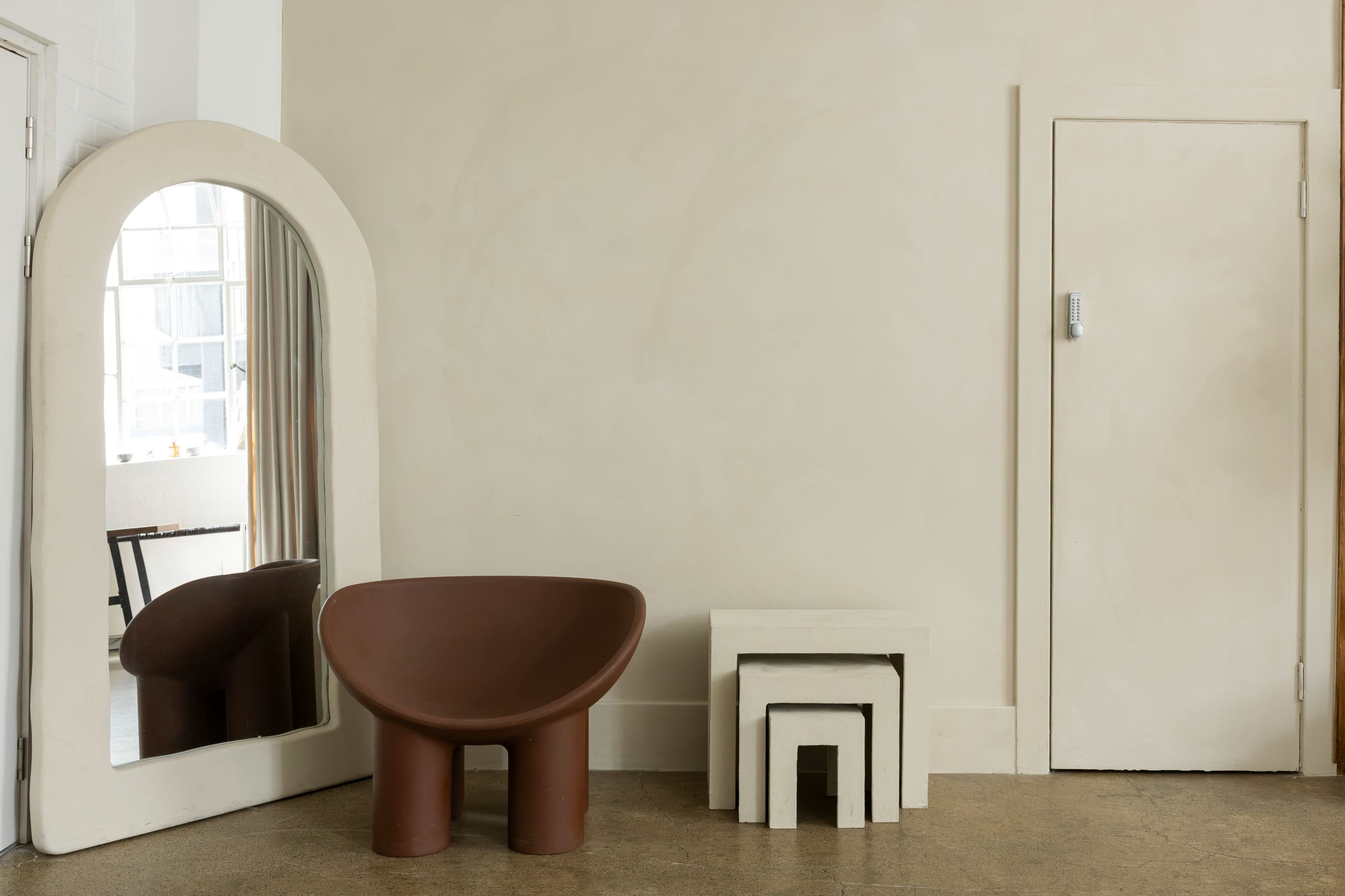 The image features a minimalist interior with a large, curved brown chair, a white nesting table set, a tall mirror with an arched top, and a door along a smooth, beige wall.