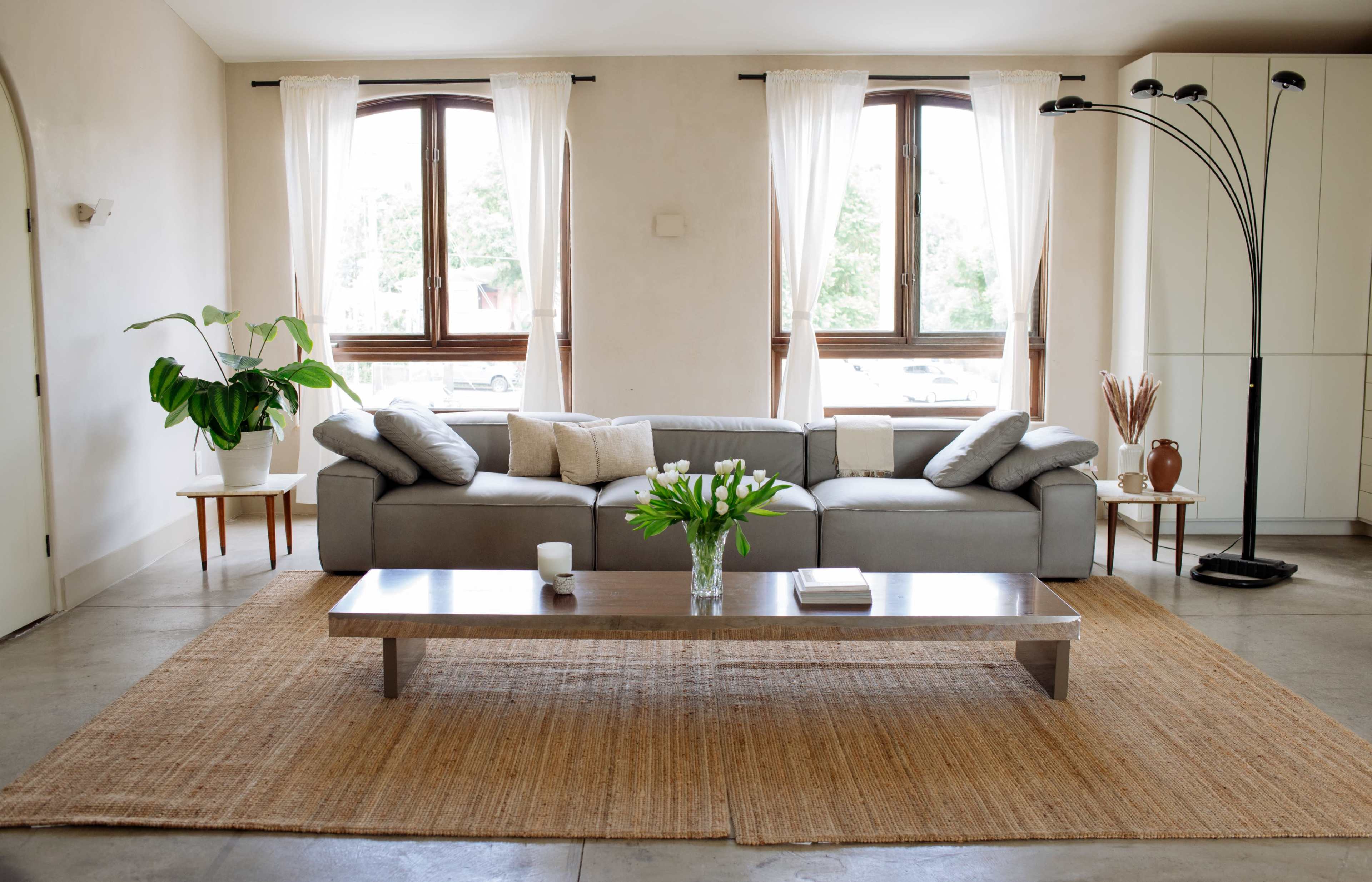 A modern living room features a gray sofa with pillows, a wooden coffee table, and large windows with sheer curtains, complemented by potted plants and a floor lamp.