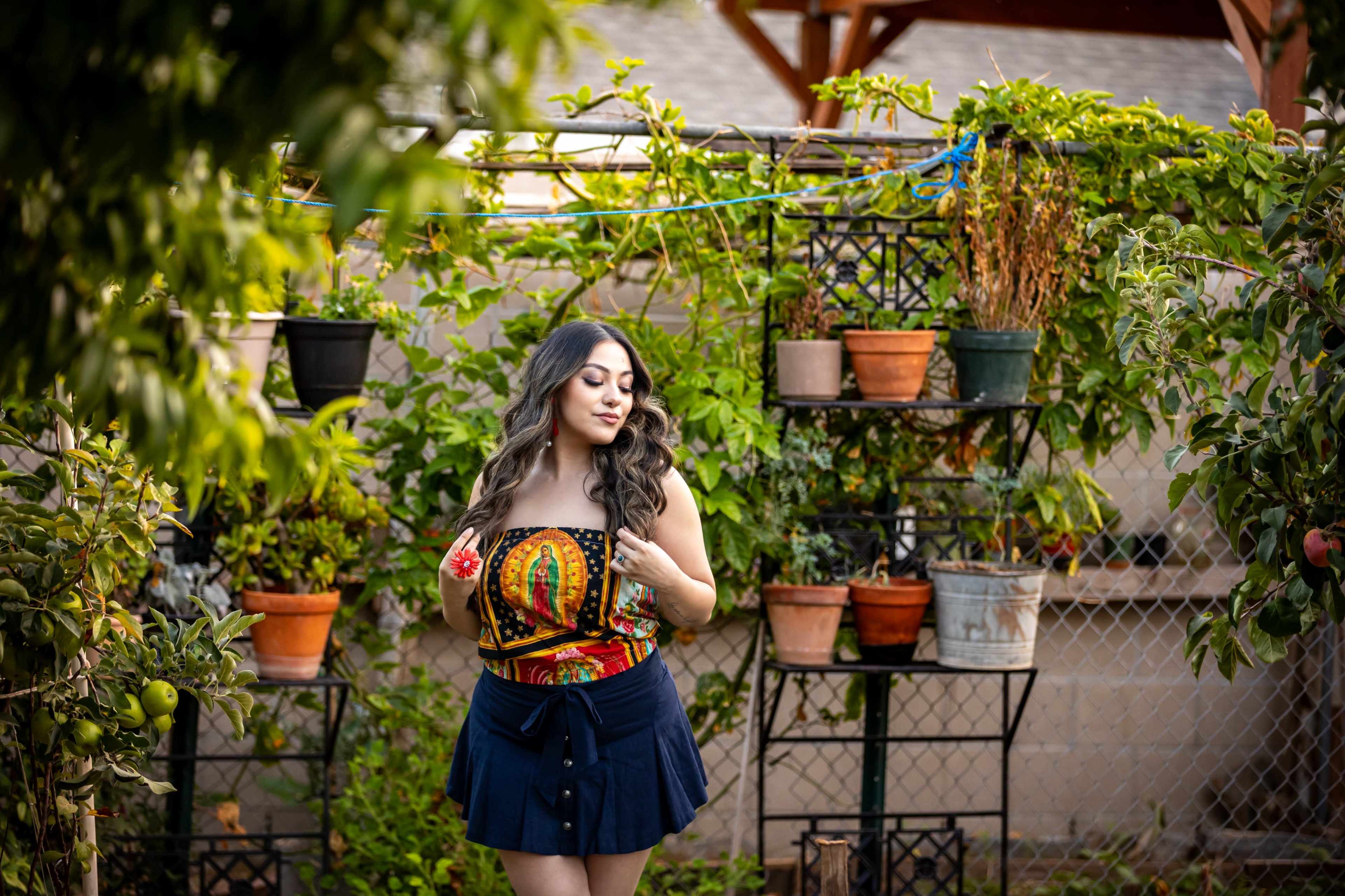 A woman stands in a garden surrounded by various potted plants and flowers.