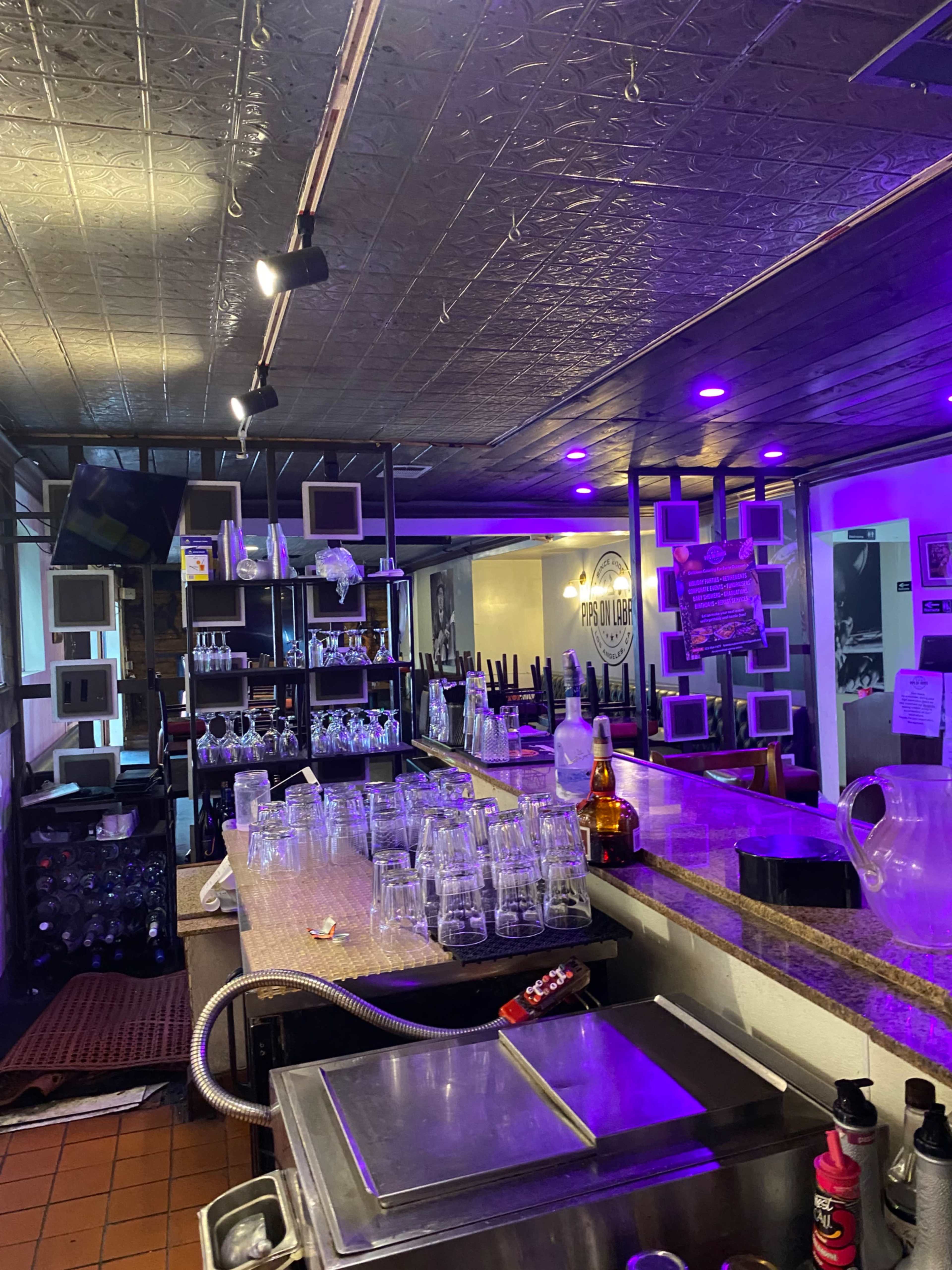 The image shows an empty bar area with a countertop lined with glassware and purple lighting illuminating the space.